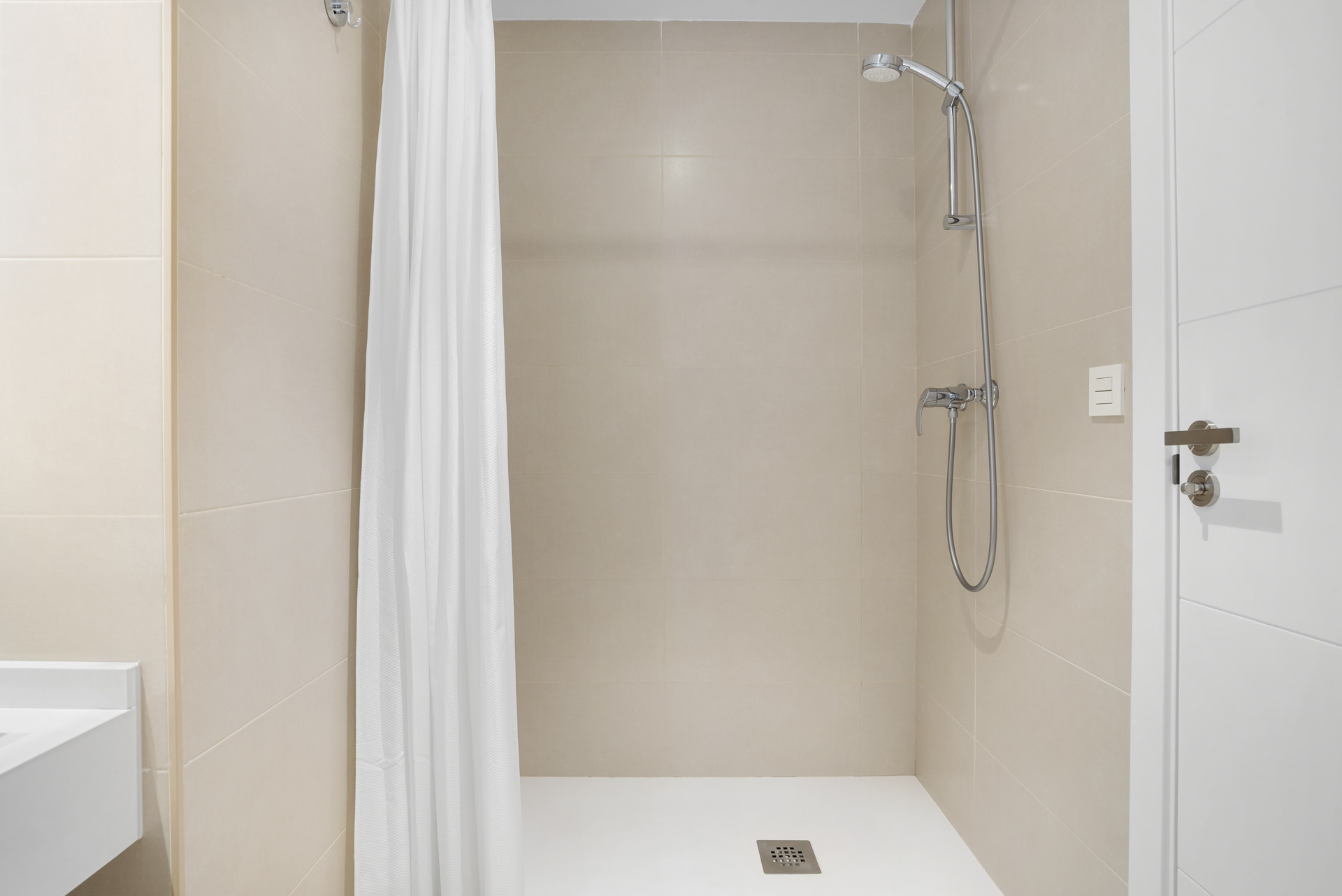 Tiled shower stall with a shower head, faucet, white curtain, and a white door partially visible.