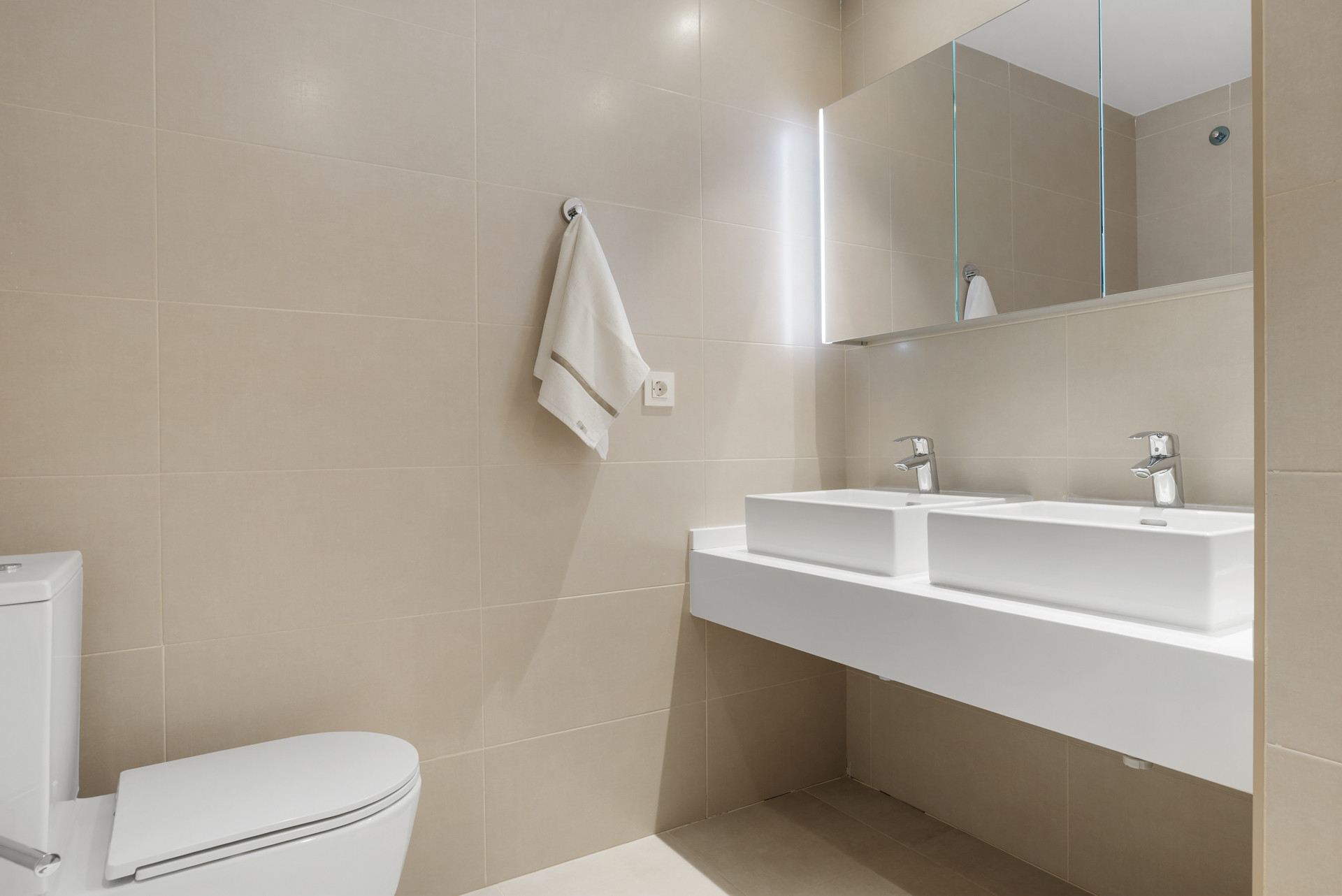 Bathroom with light beige tiles, dual rectangular sinks, a toilet, and a large mirrored cabinet.