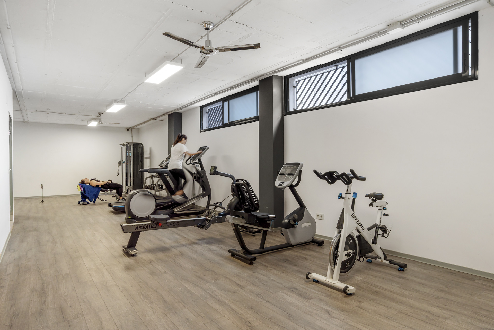Indoor gym with various exercise machines, two people working out, and ceiling fans.