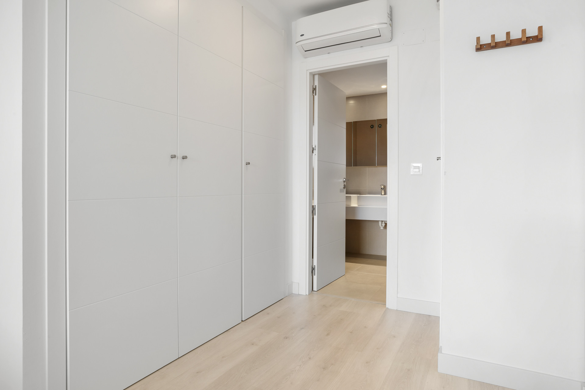 White hallway with built-in wardrobes, a coat rack, and an open door to a bathroom with a sink.