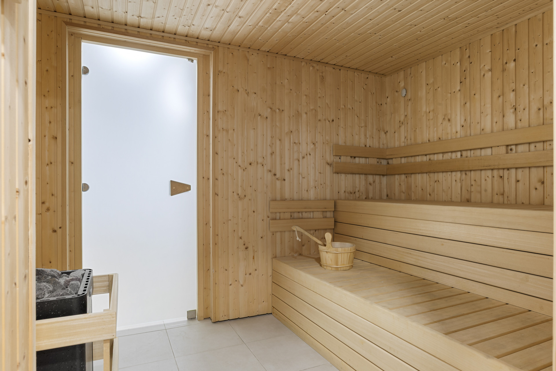 Wooden sauna with slatted benches, a bucket and ladle, and a glass door leading out.
