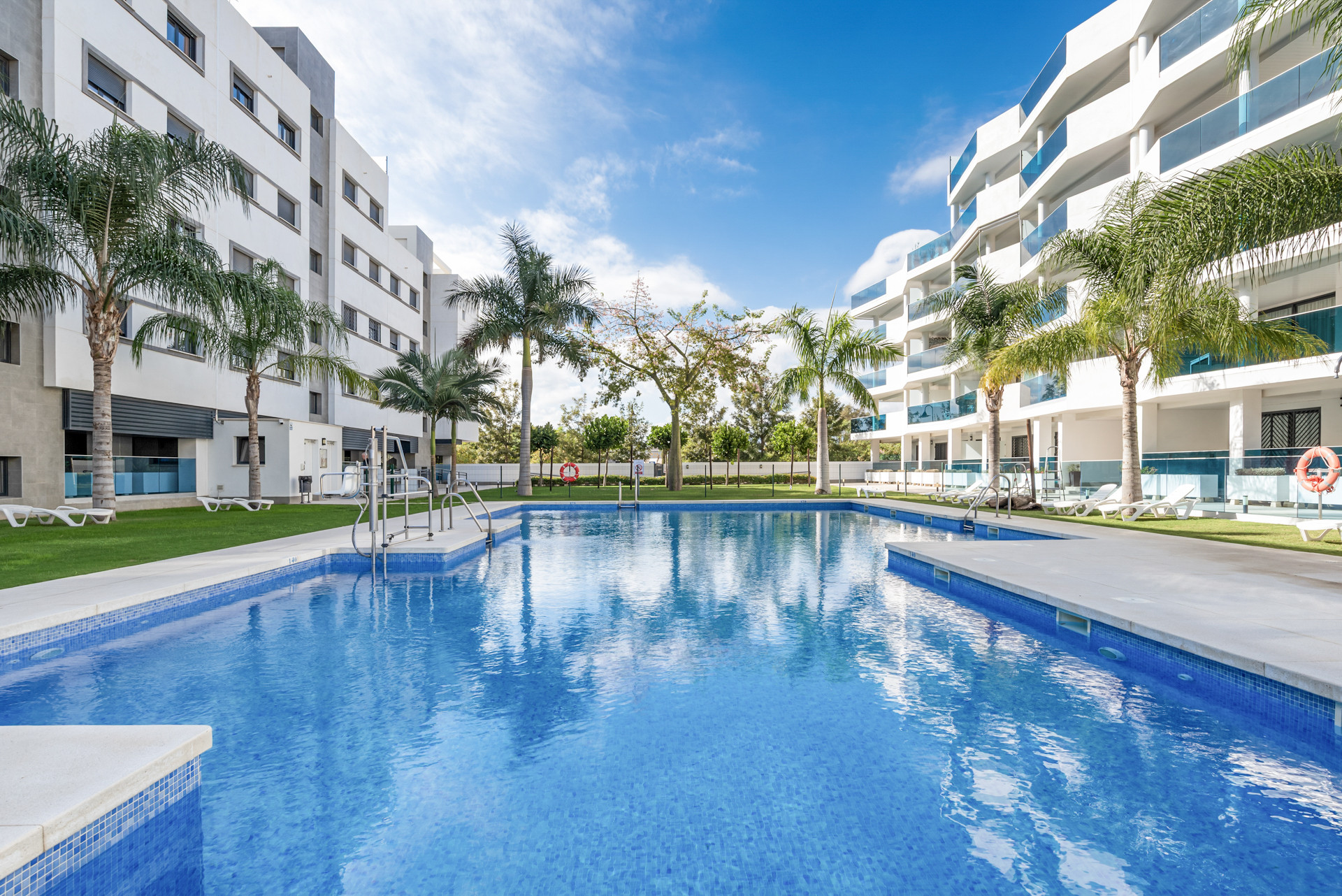 Outdoor view of a large swimming pool with surrounding palm trees and modern apartment buildings under a blue sky.