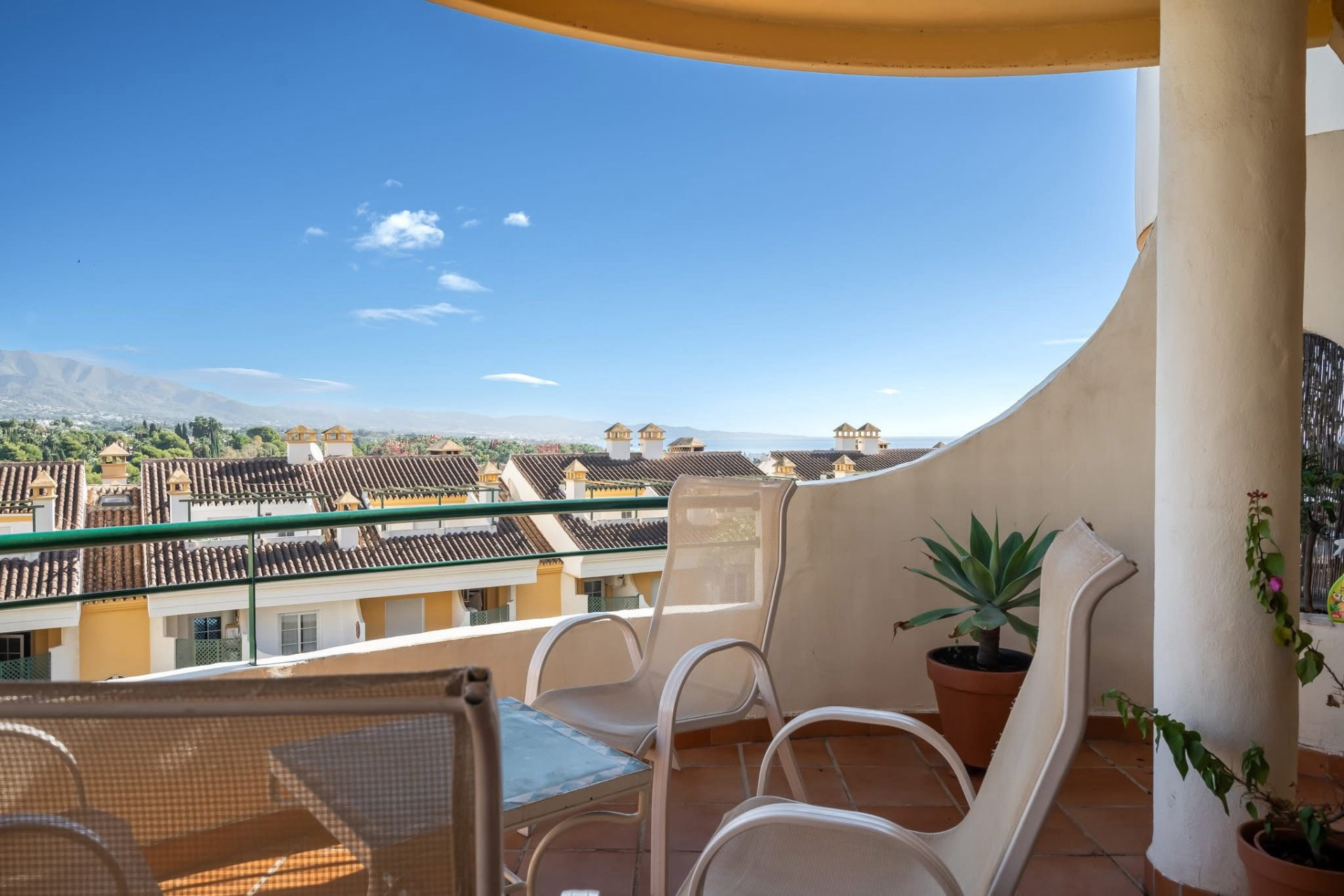 Balcony with two lounge chairs and potted plant, overlooking red-tiled roofs and distant ocean.