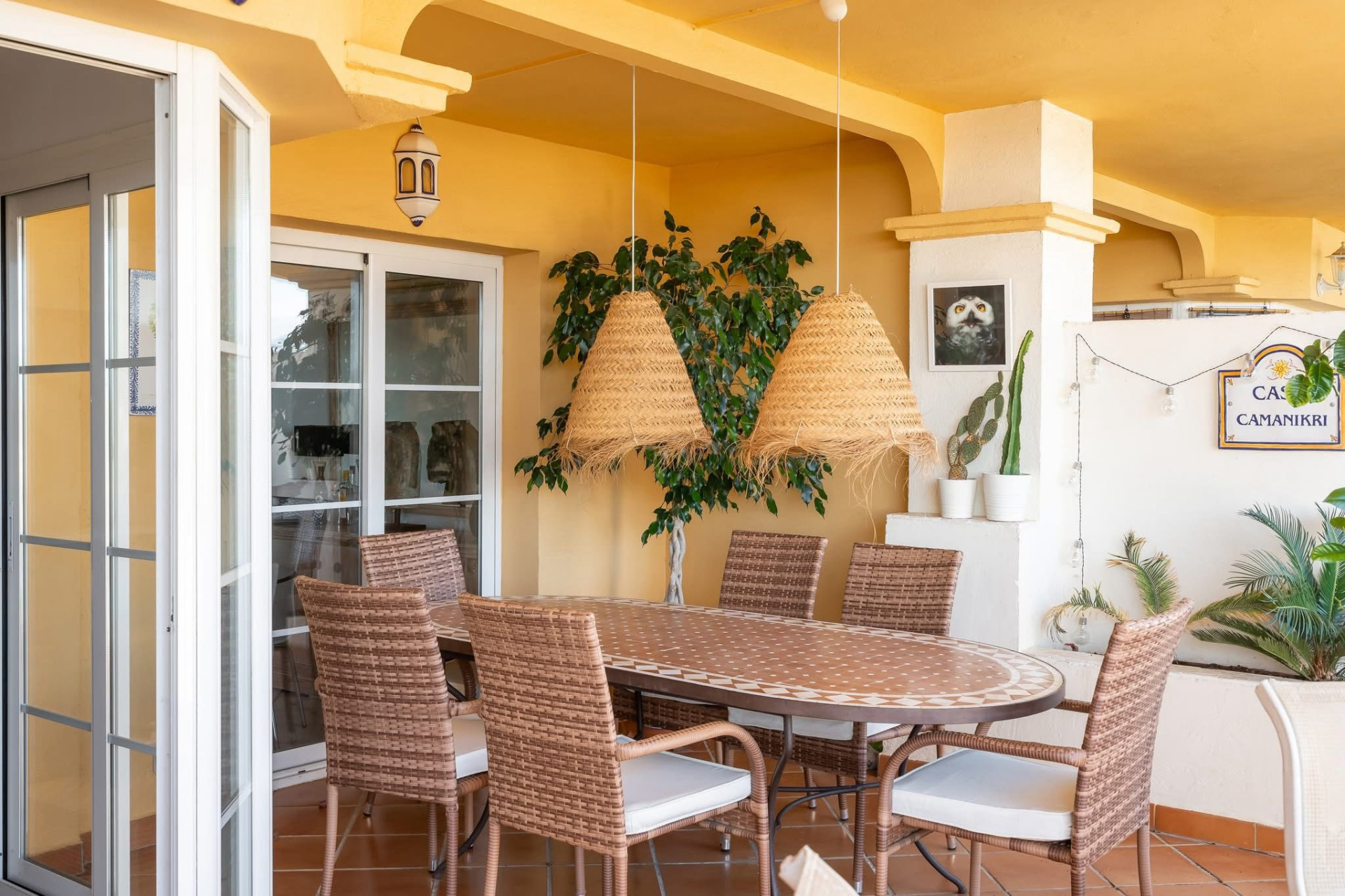 Outdoor patio with a large dining table, wicker chairs, yellow walls, and hanging straw light fixtures.