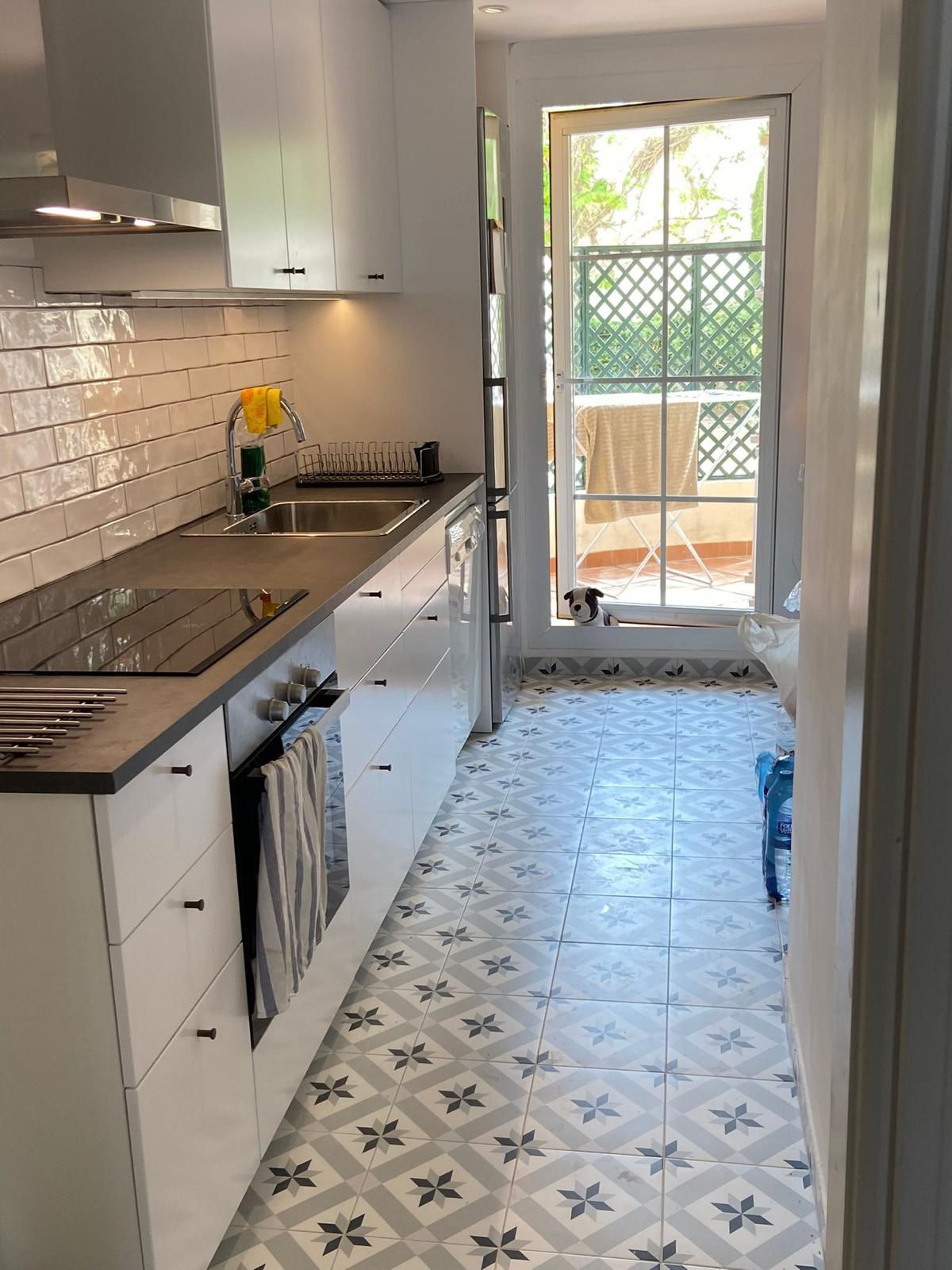 Galley kitchen with white cabinets, dark countertops, patterned floor tiles, and a small dog peeking from the patio.
