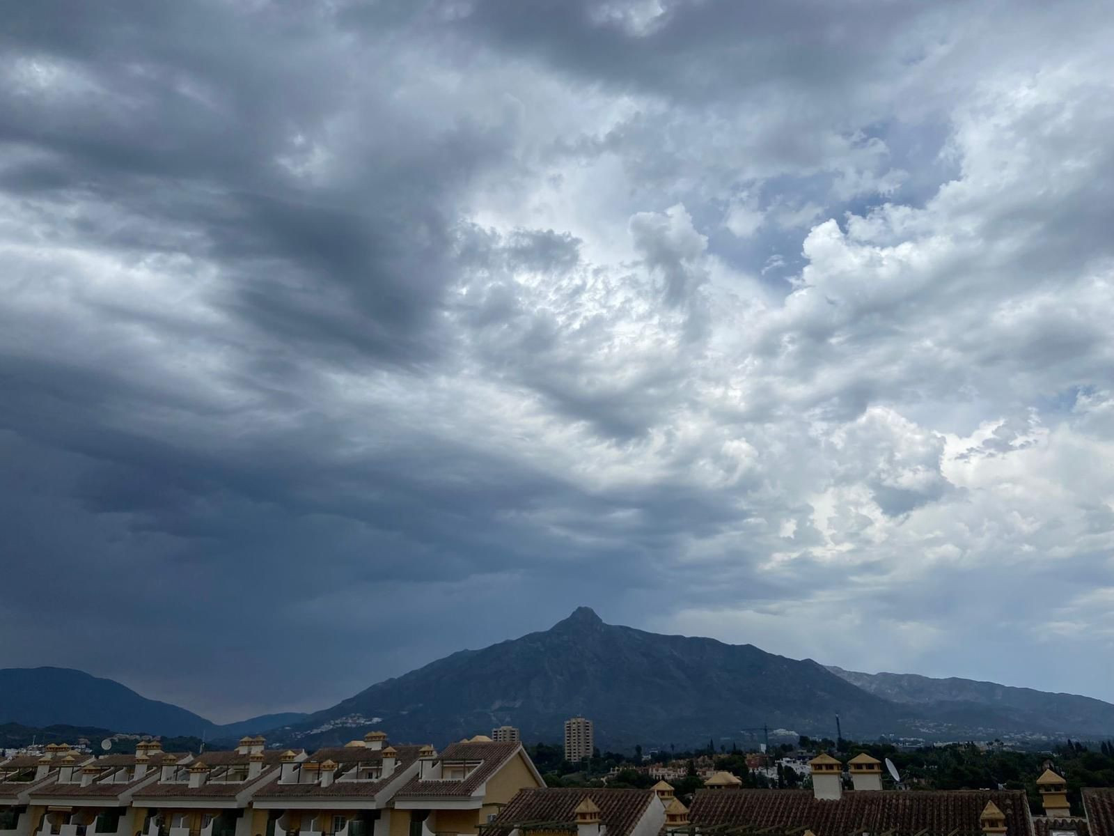 View of a large mountain under a dramatic, cloudy sky, with building rooftops in the foreground.