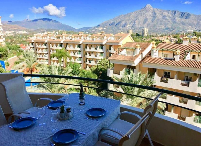 Balcony with a set dining table, overlooking buildings, palm trees, and mountains under a blue sky.