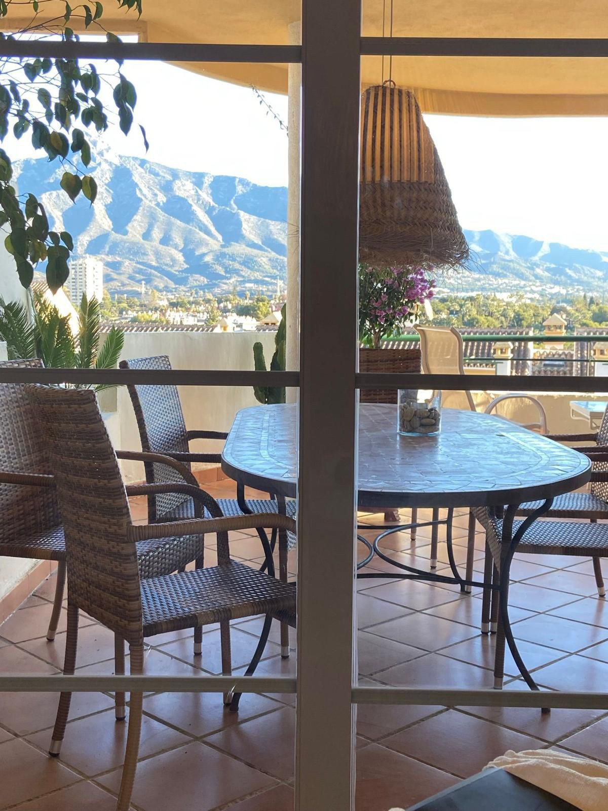Terrace with a mosaic dining table and woven chairs, seen through glass doors, mountain view.