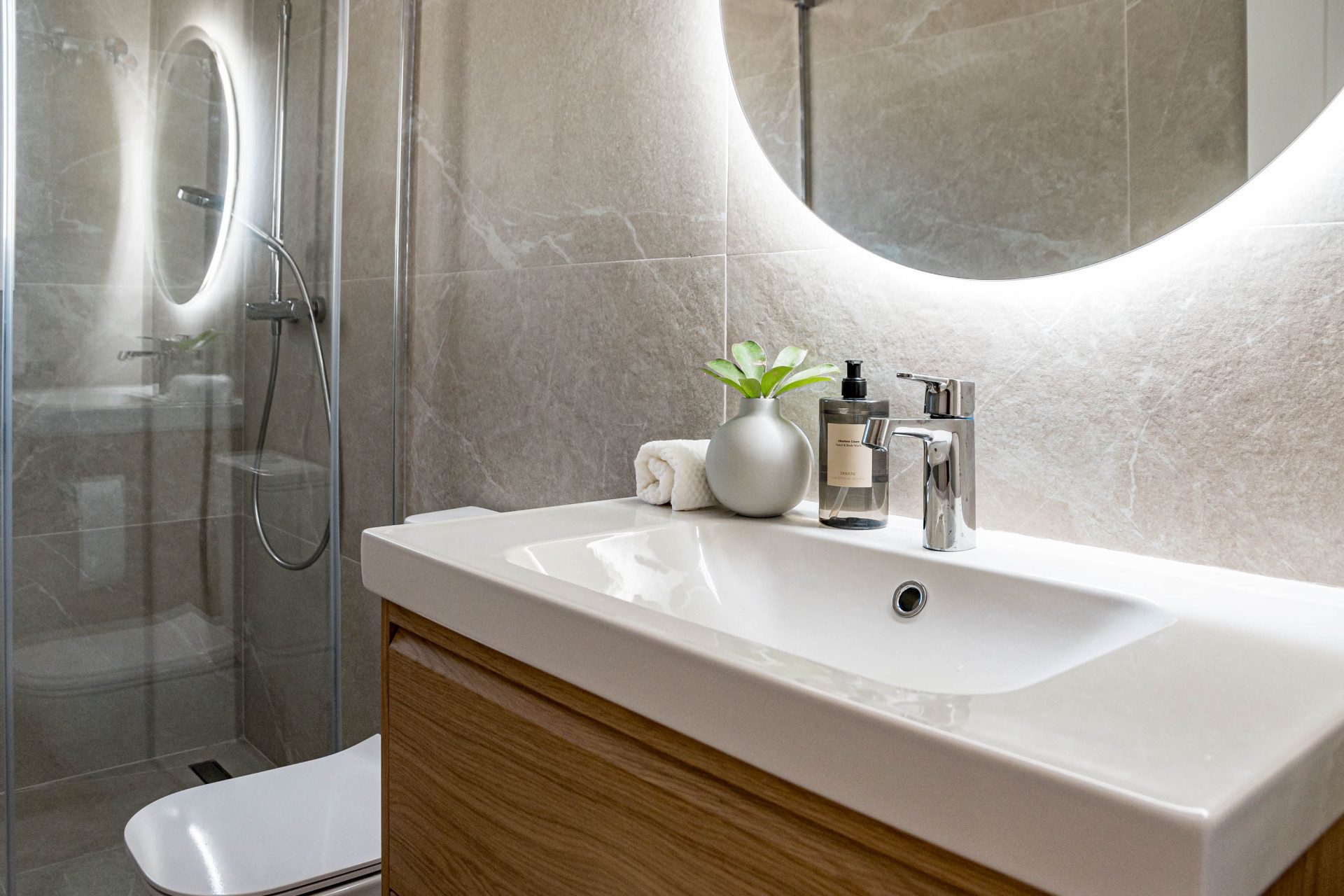 Close up of bathroom sink with wooden vanity, chrome faucet, small plant, and illuminated mirror.