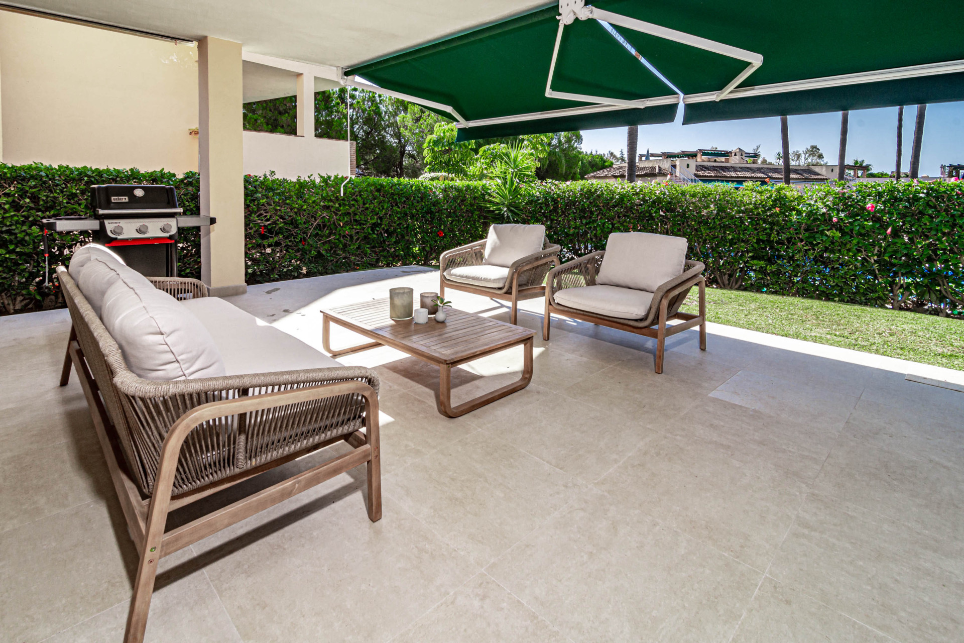 Outdoor patio with a barbecue grill, three outdoor seating pieces, and a large green hedge in the background.