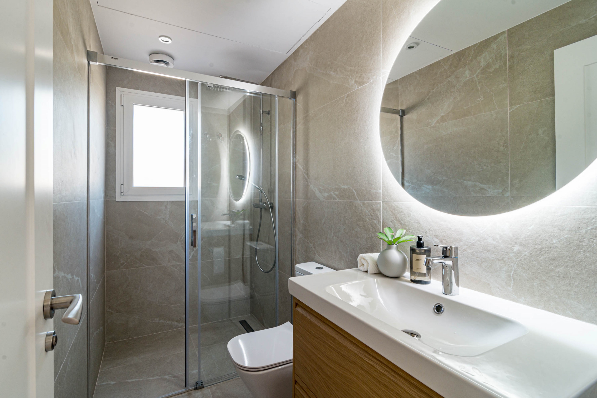 Modern bathroom with walk-in shower, wooden vanity, sink, toilet, and backlit round mirror.