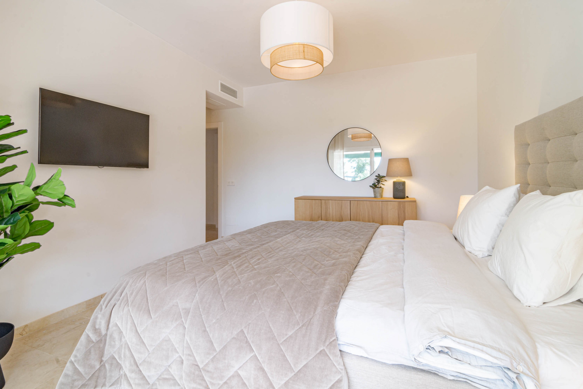 Bedroom view towards a wooden console table with a round mirror, and a wall-mounted TV.