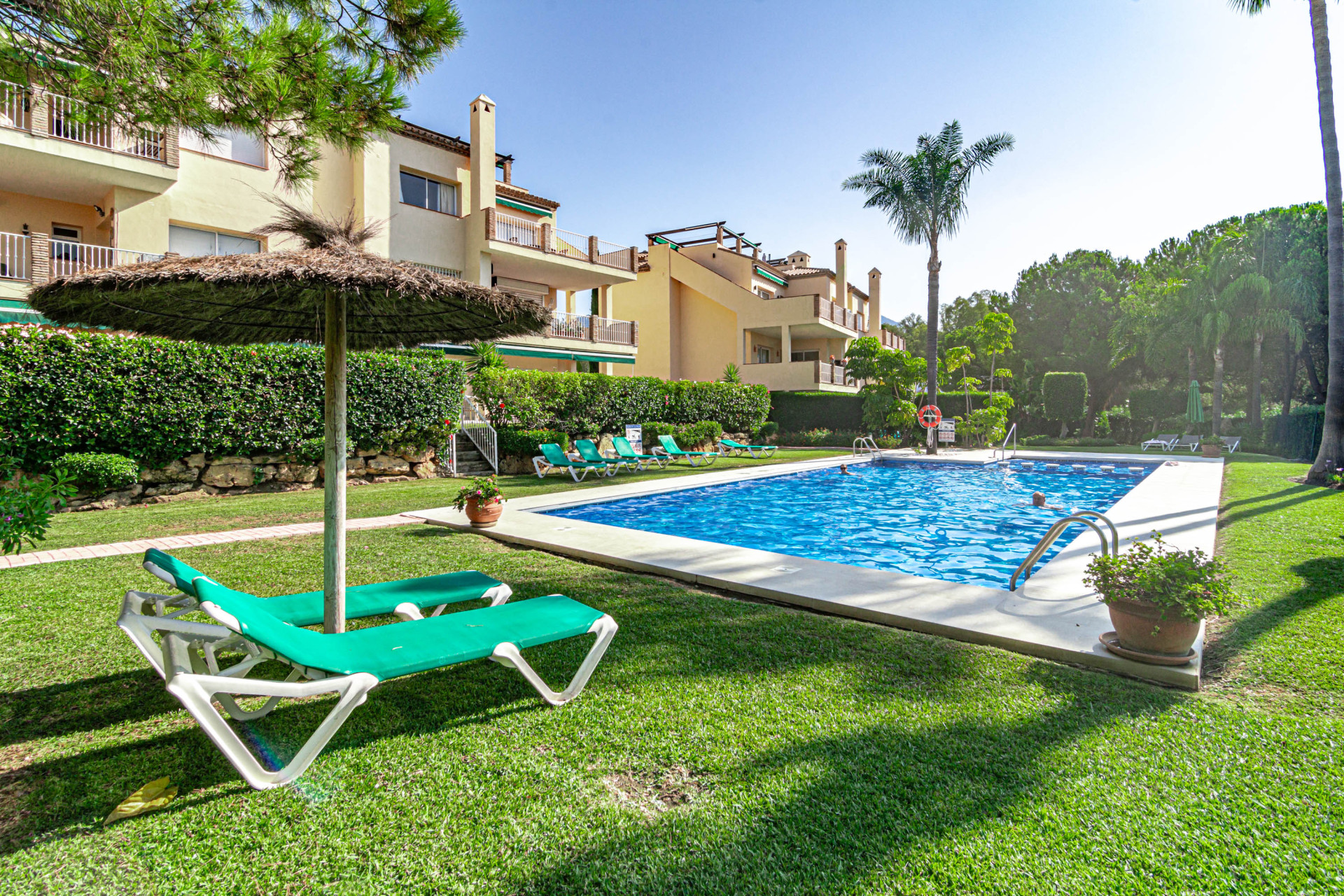 Outdoor pool area with green grass, sun loungers, umbrella, and apartment buildings under blue sky.