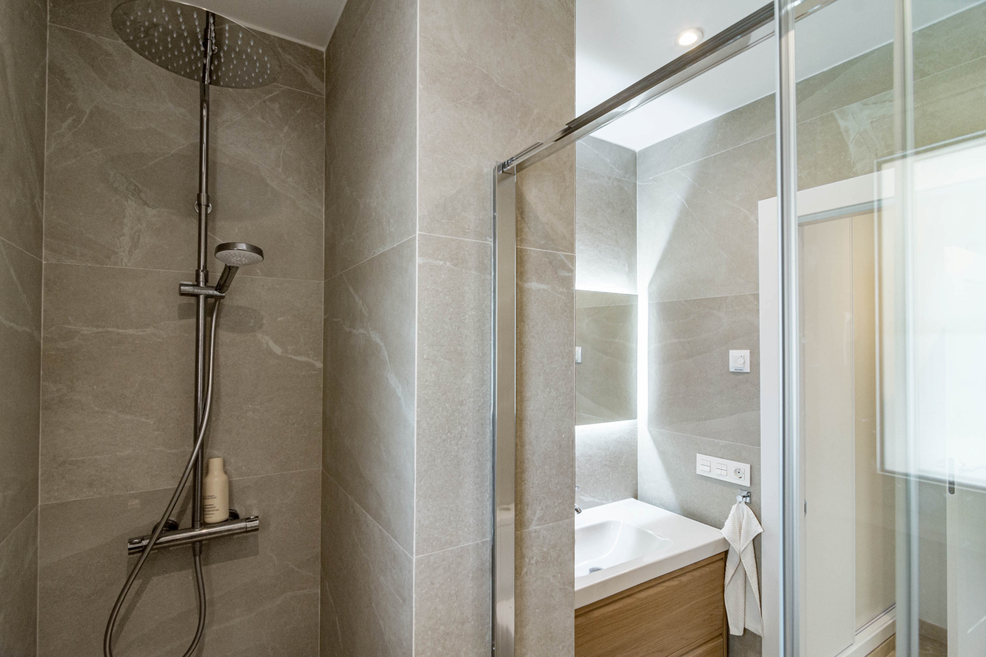 Shower area with light gray tiled walls, a rain showerhead, and a glass partition.