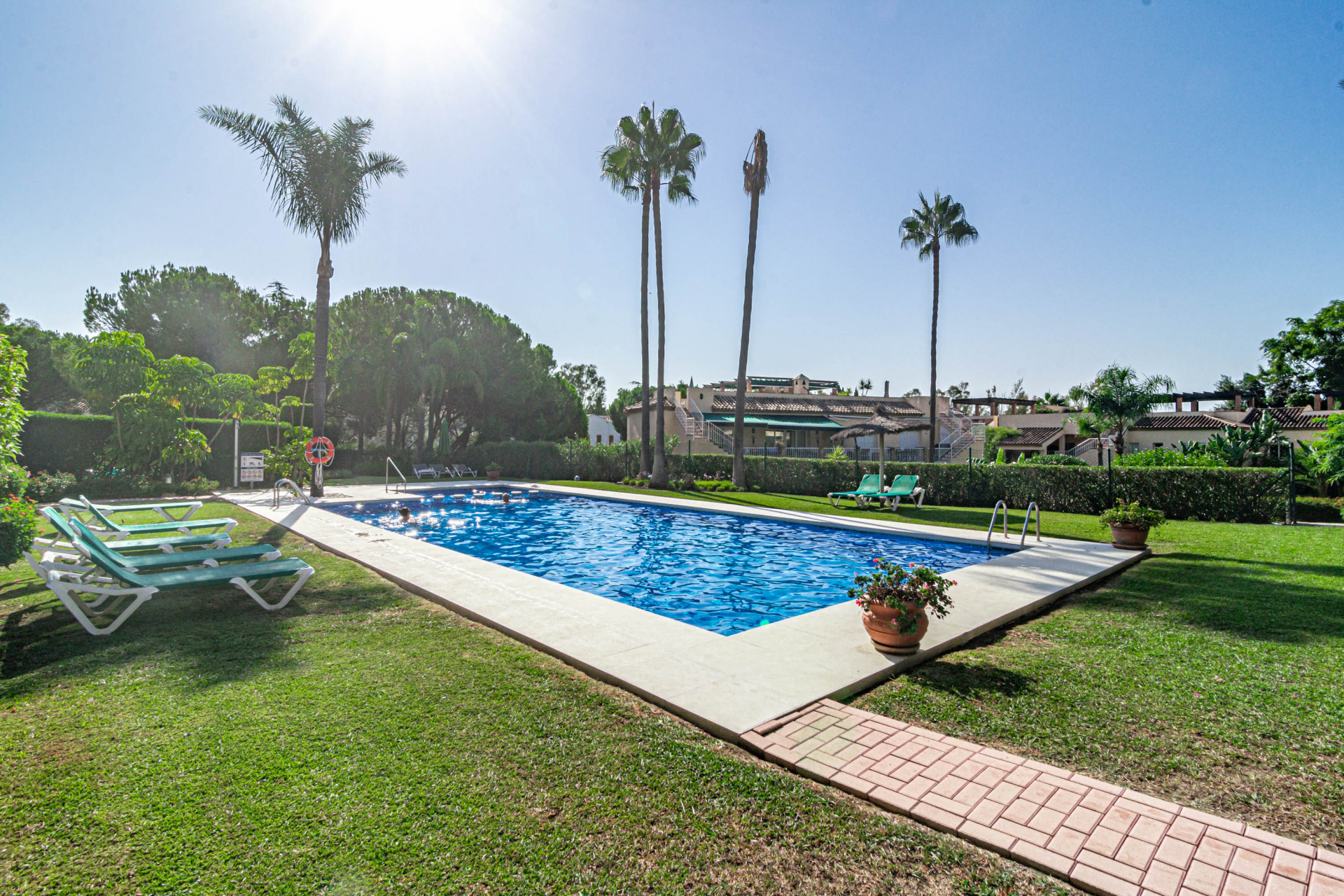 Expansive view of a community pool area with a red brick path, palm trees, and green lawns.