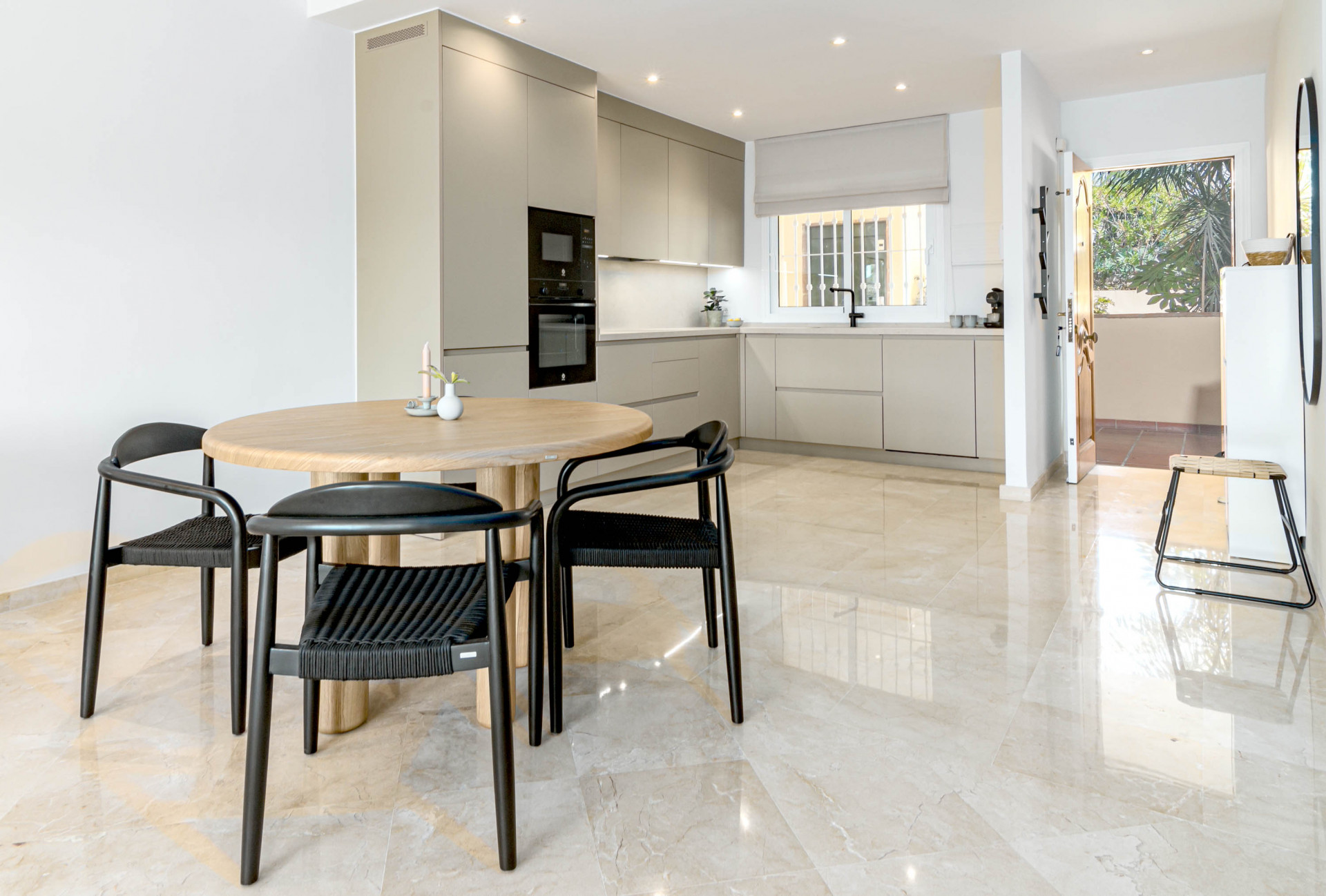 Open plan kitchen with light wood dining table and black chairs on marble floor, facing an outdoor patio.