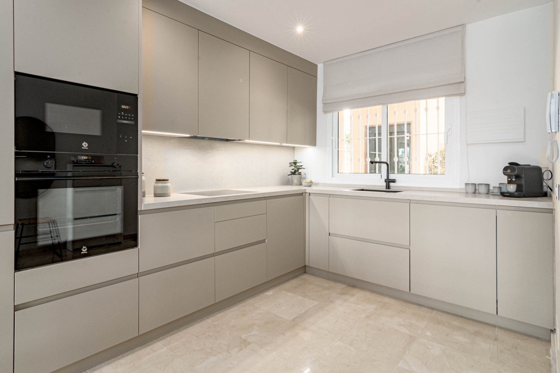 Modern kitchen with built-in oven, light grey cabinetry, black sink, and a barred window.