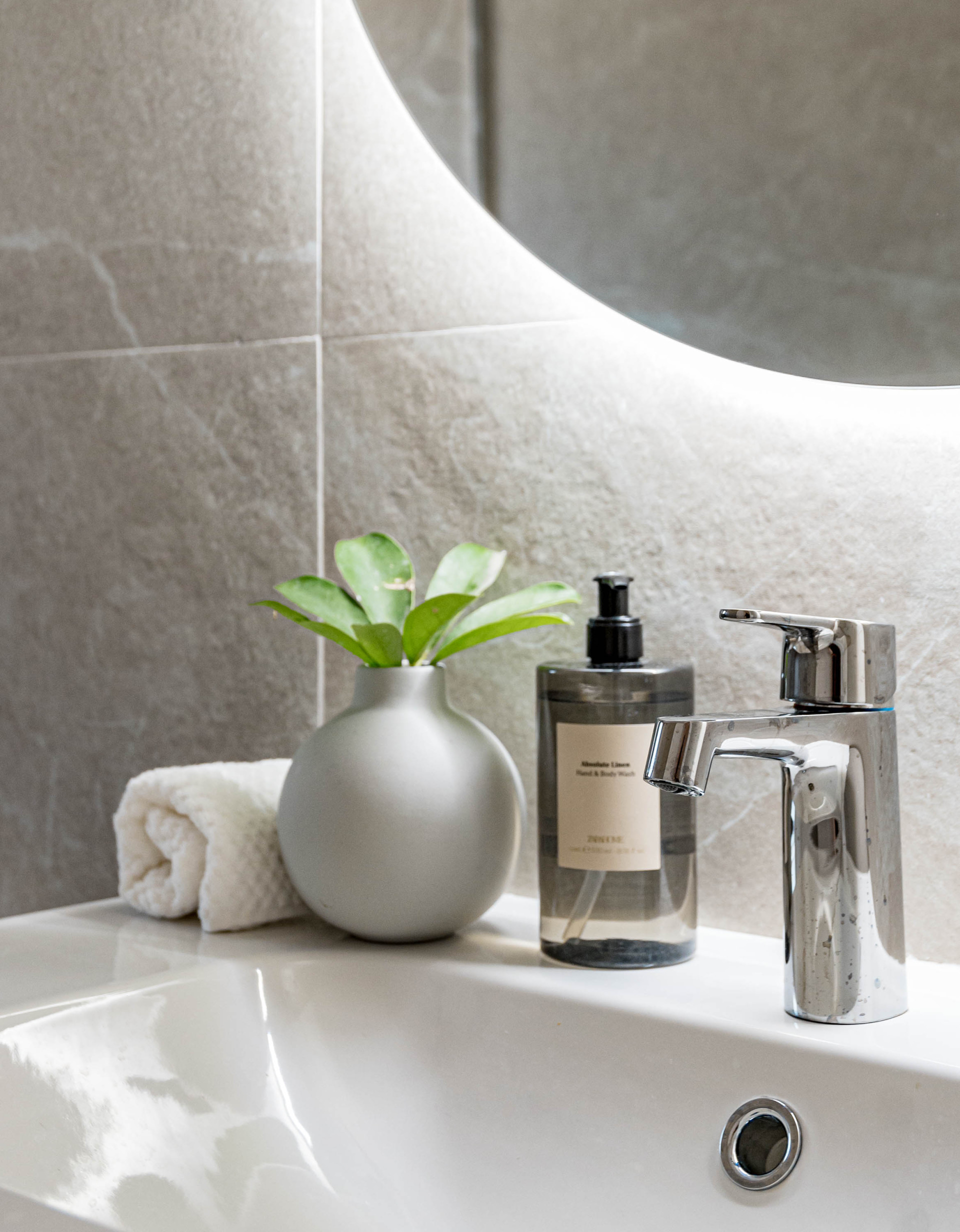 Close-up of a white bathroom sink with a chrome faucet, a grey tiled wall, and a backlit round mirror.