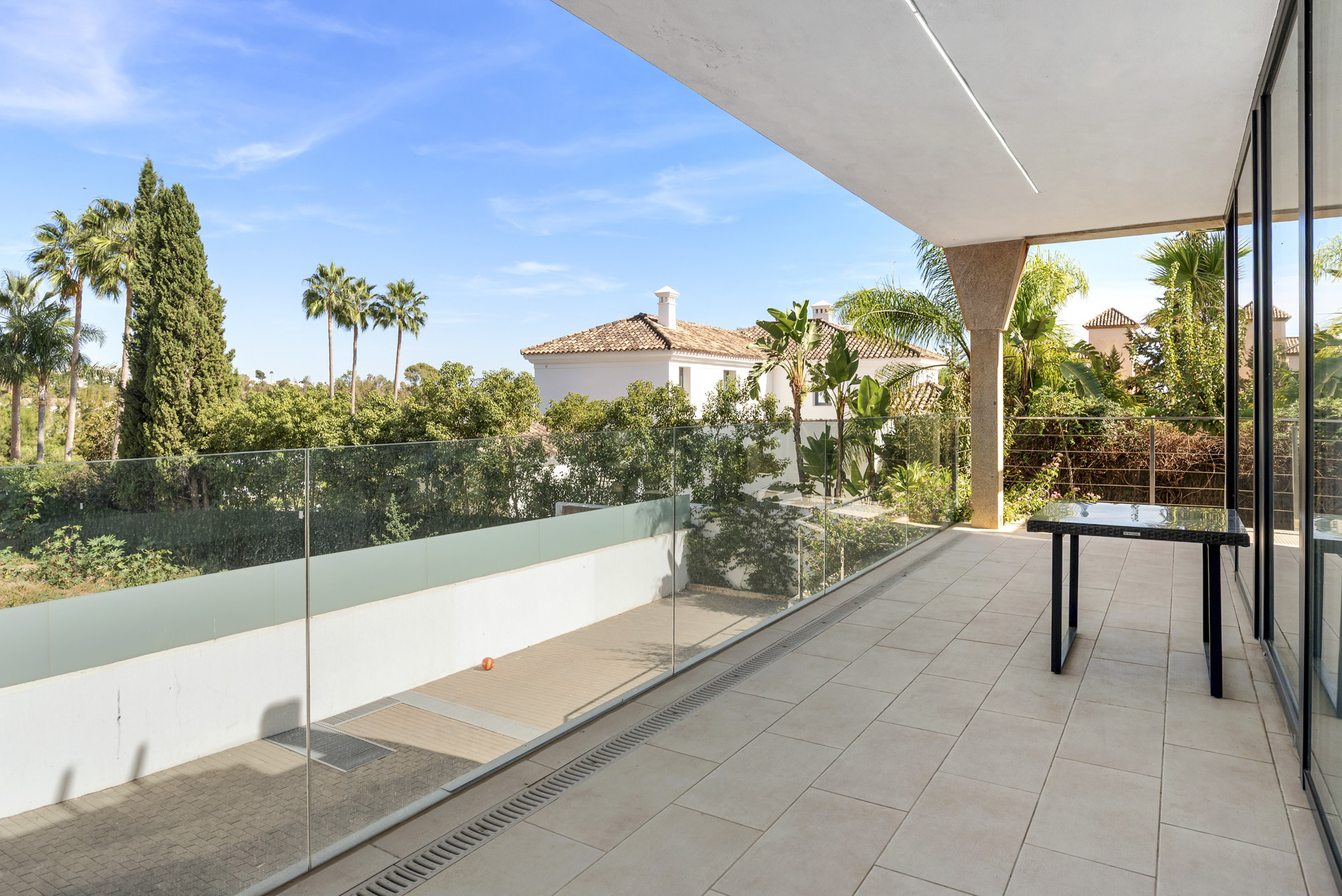 Balcony view with glass railing overlooking greenery and palm trees under a clear blue sky.