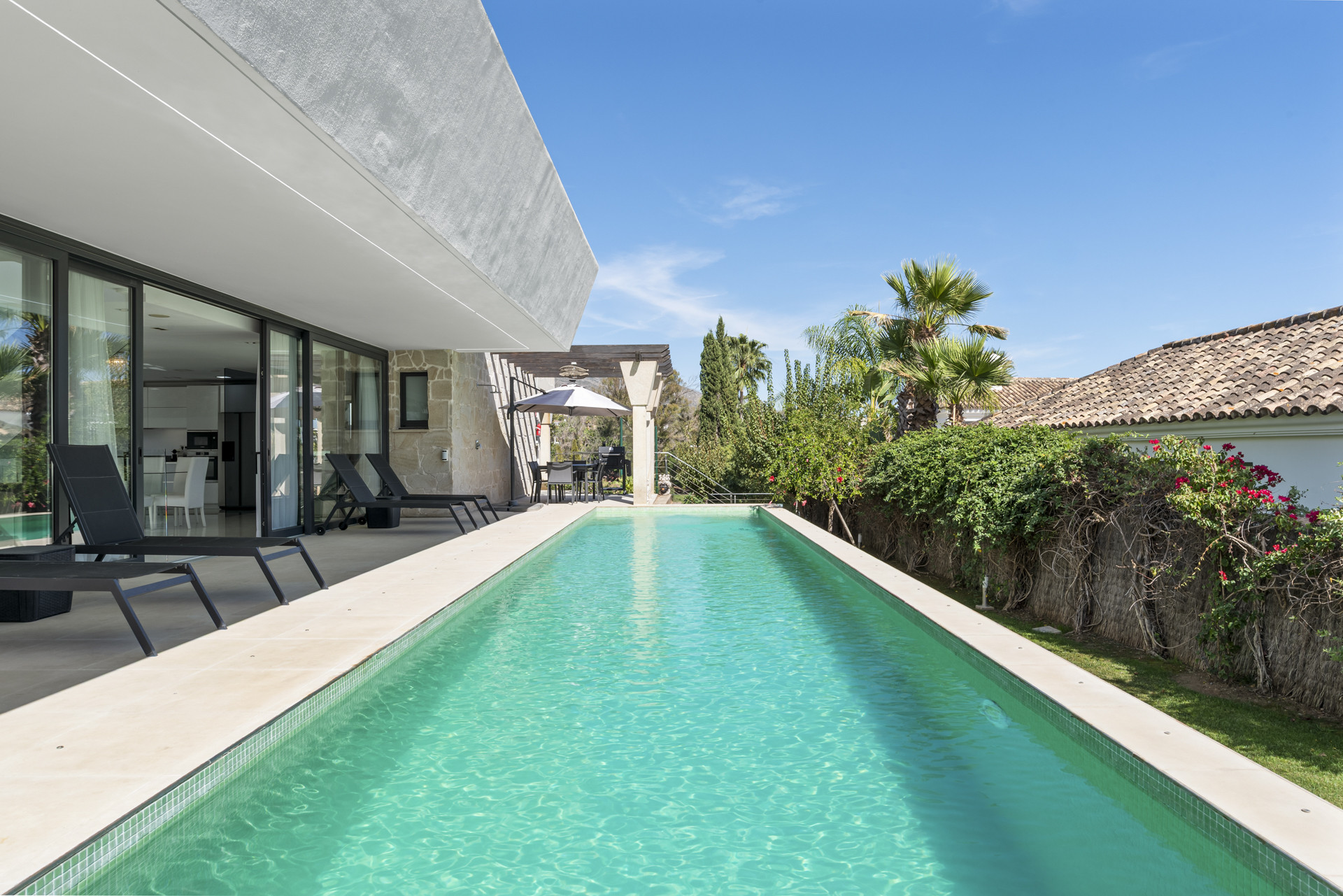Swimming pool bordered by lounge chairs, visible from the garden perspective against the blue sky.
