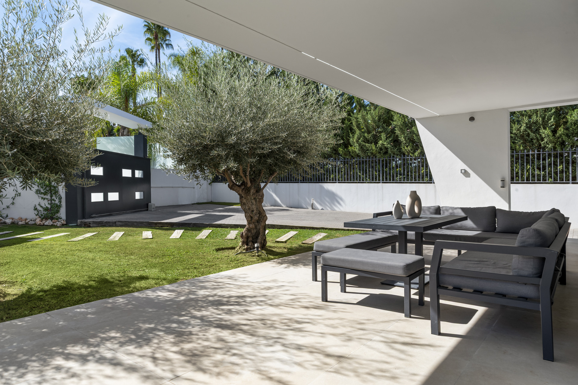Covered patio with seating area and olive tree in a garden setting, showcasing modern outdoor design.
