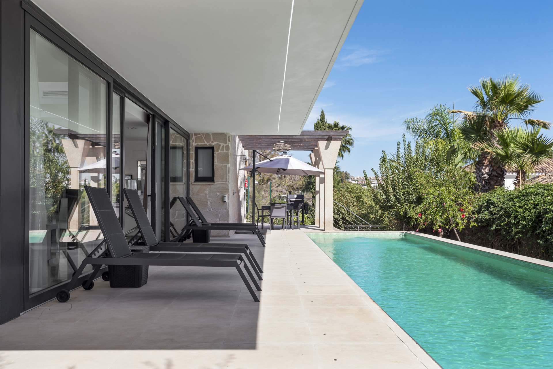 Lounge area outside with sun loungers beside a swimming pool and dining table under an umbrella.