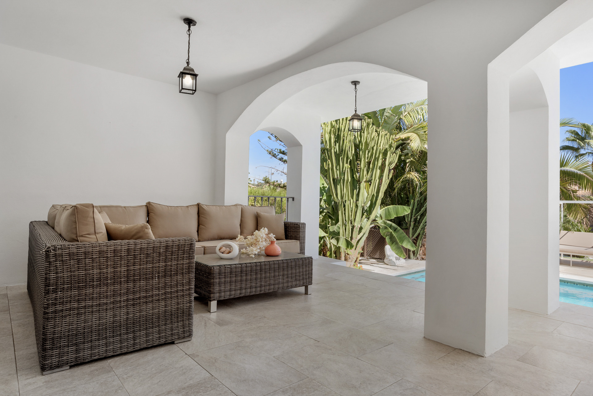 Covered patio with two L-shaped wicker sofas, a coffee table, and an archway to lush foliage.