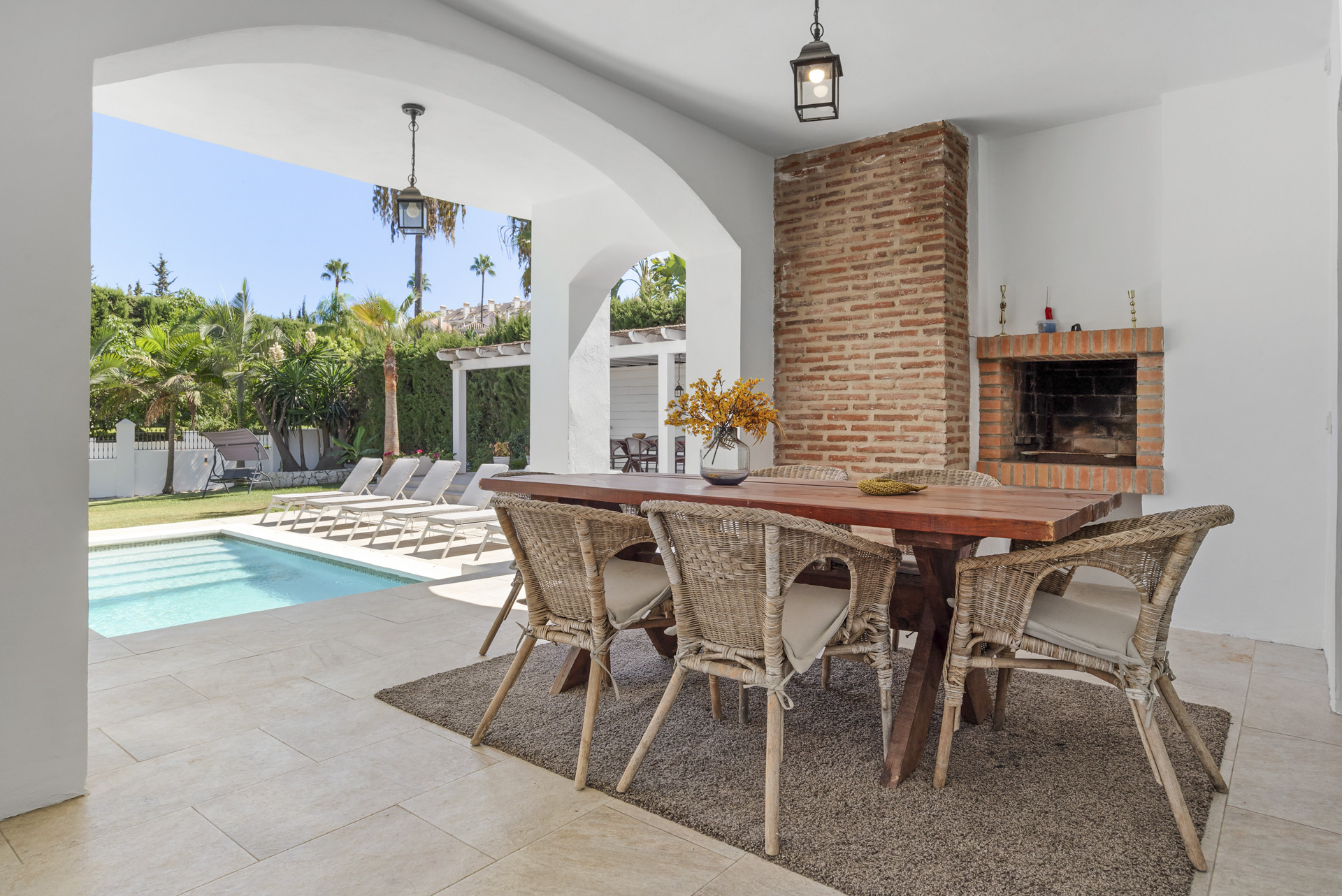 Covered outdoor dining area with a brick BBQ and view of a swimming pool and loungers.