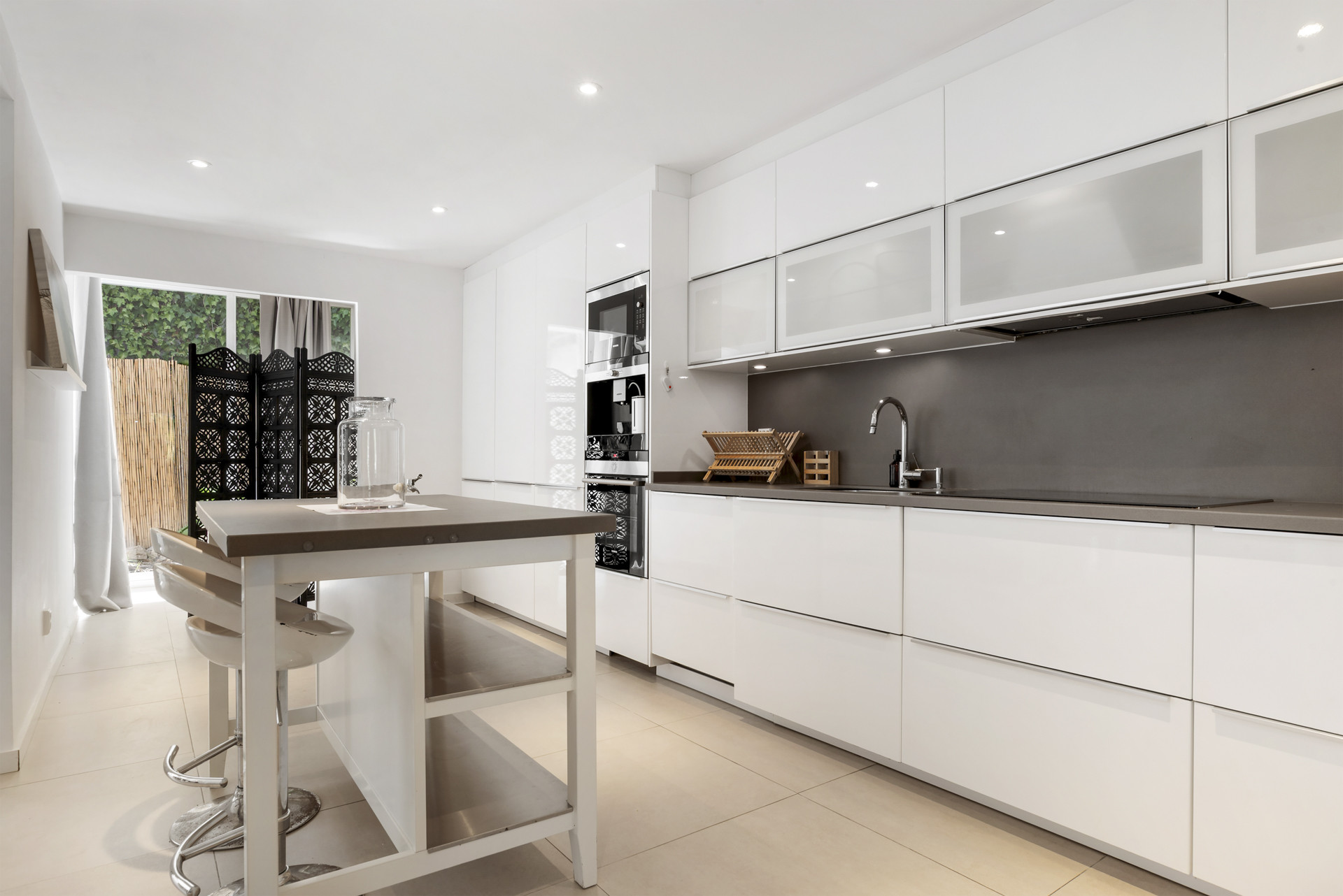 Sleek white kitchen featuring a central island with seating and a view of outdoor greenery through a window.