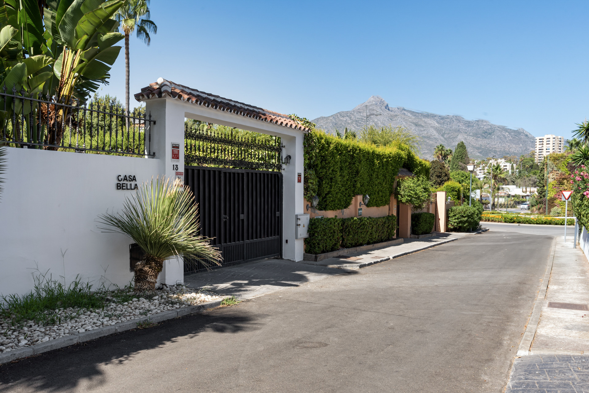 Street view of a white wall with a black gate, tropical plants, and a distant mountain.