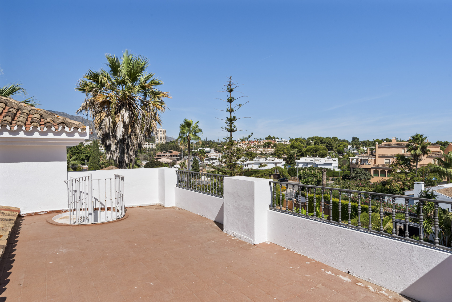 Rooftop terrace with red tiled flooring, white perimeter walls, and distant city views.