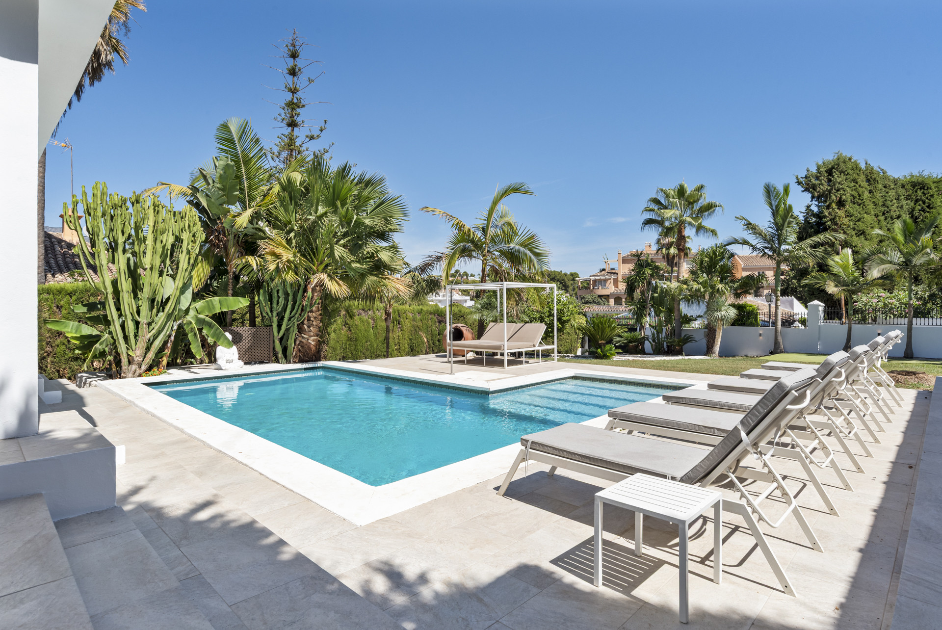 A view of the rectangular swimming pool and surrounding patio with multiple sun loungers and lush greenery.