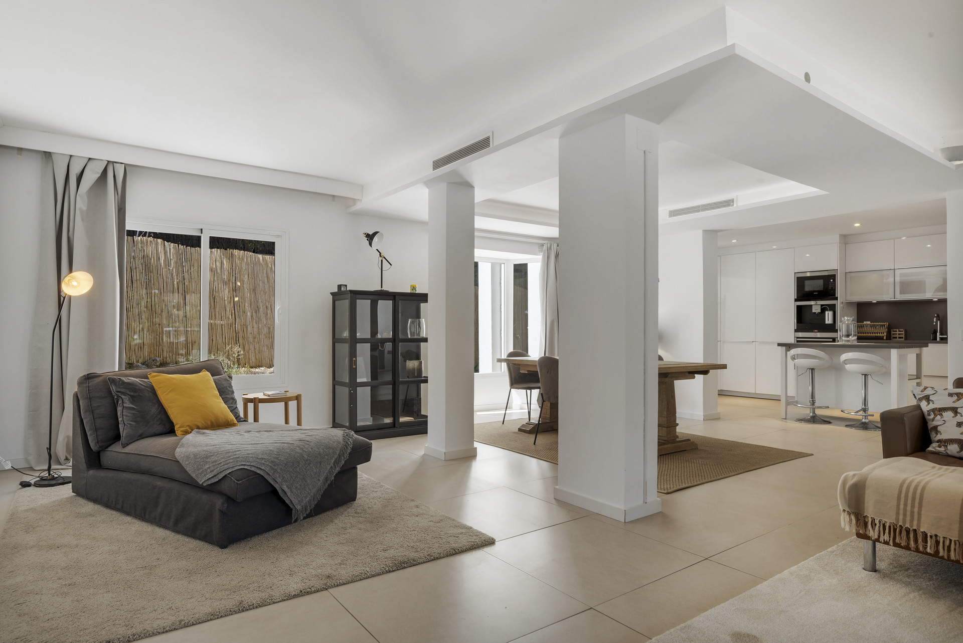 Open concept living space showing a gray chaise lounge, white pillars, and a distant view of the kitchen.