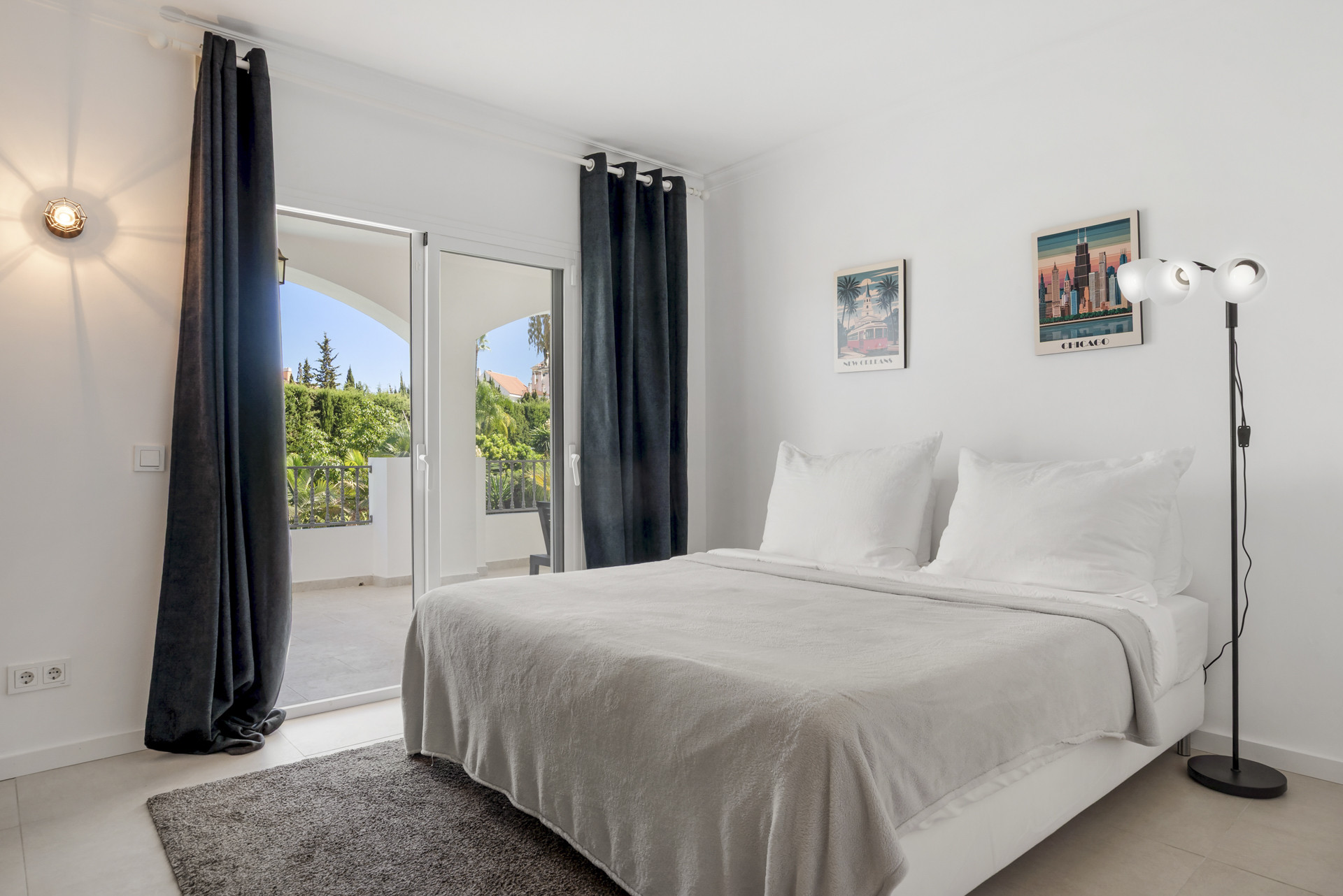 Bedroom with a white bed, dark curtains, two posters, and a glass door opening to a balcony.