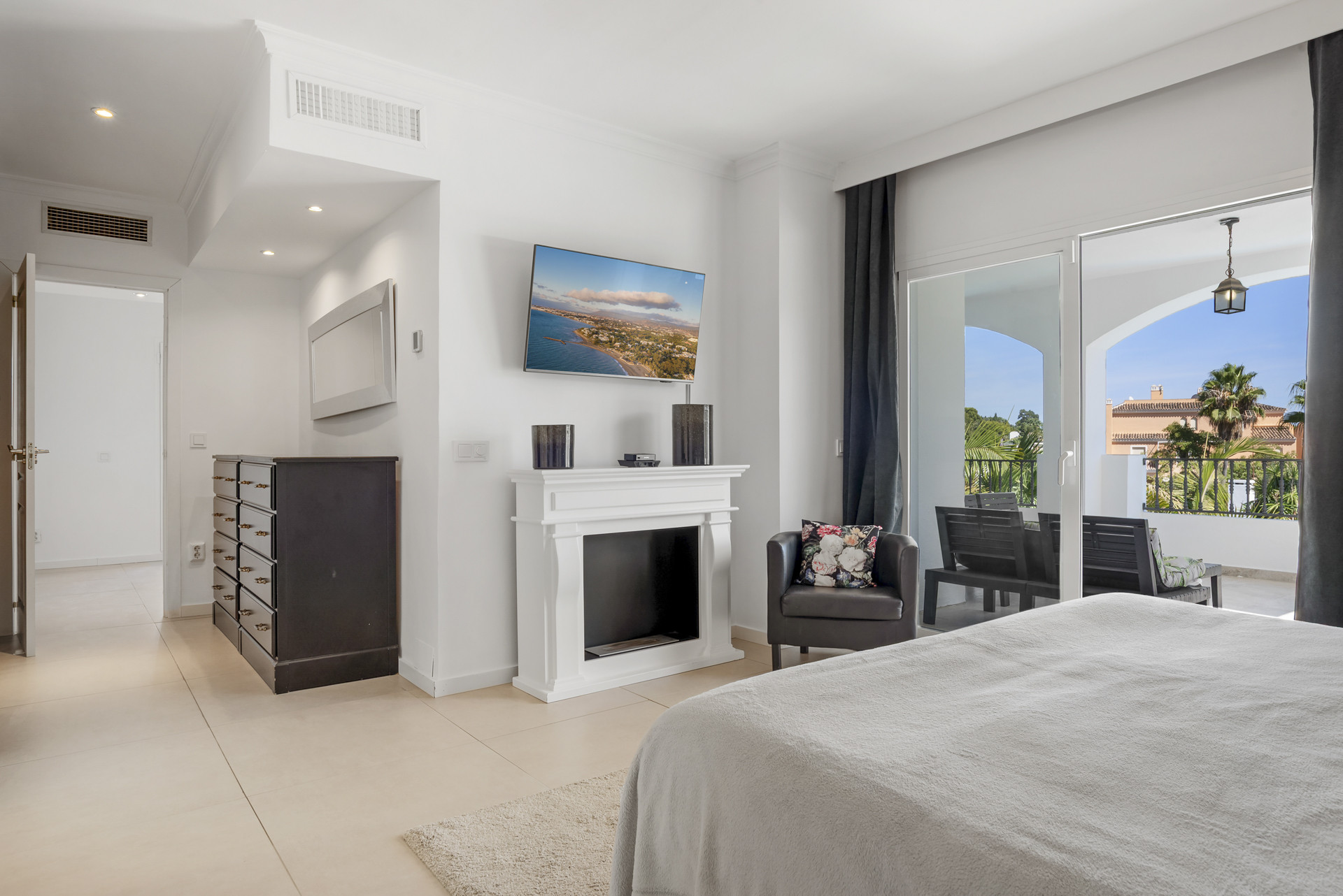 Bedroom with a large bed, wall-mounted TV above a decorative fireplace, and sliding glass doors to a balcony.
