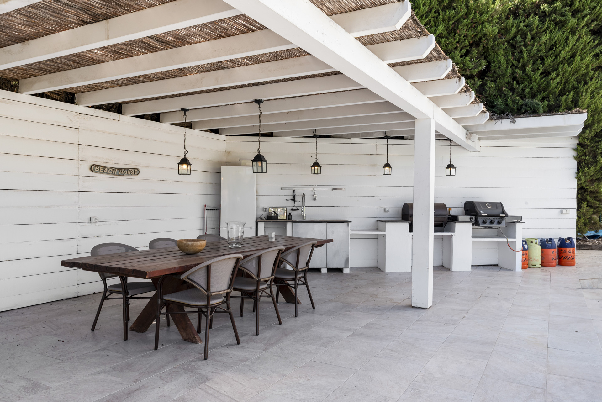 Covered outdoor kitchen and dining area with two grills, sink, and "BEACH HOUSE" sign.