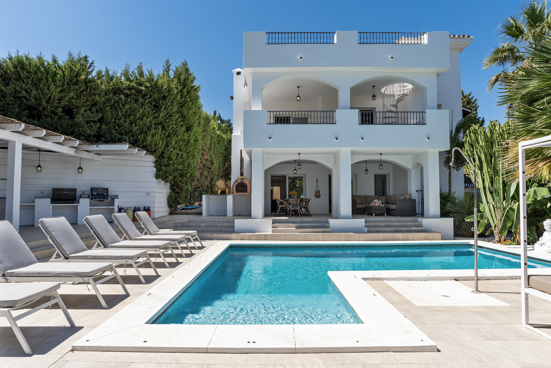 House exterior with pool in foreground, surrounded by loungers, lush hedges, and an outdoor dining area.