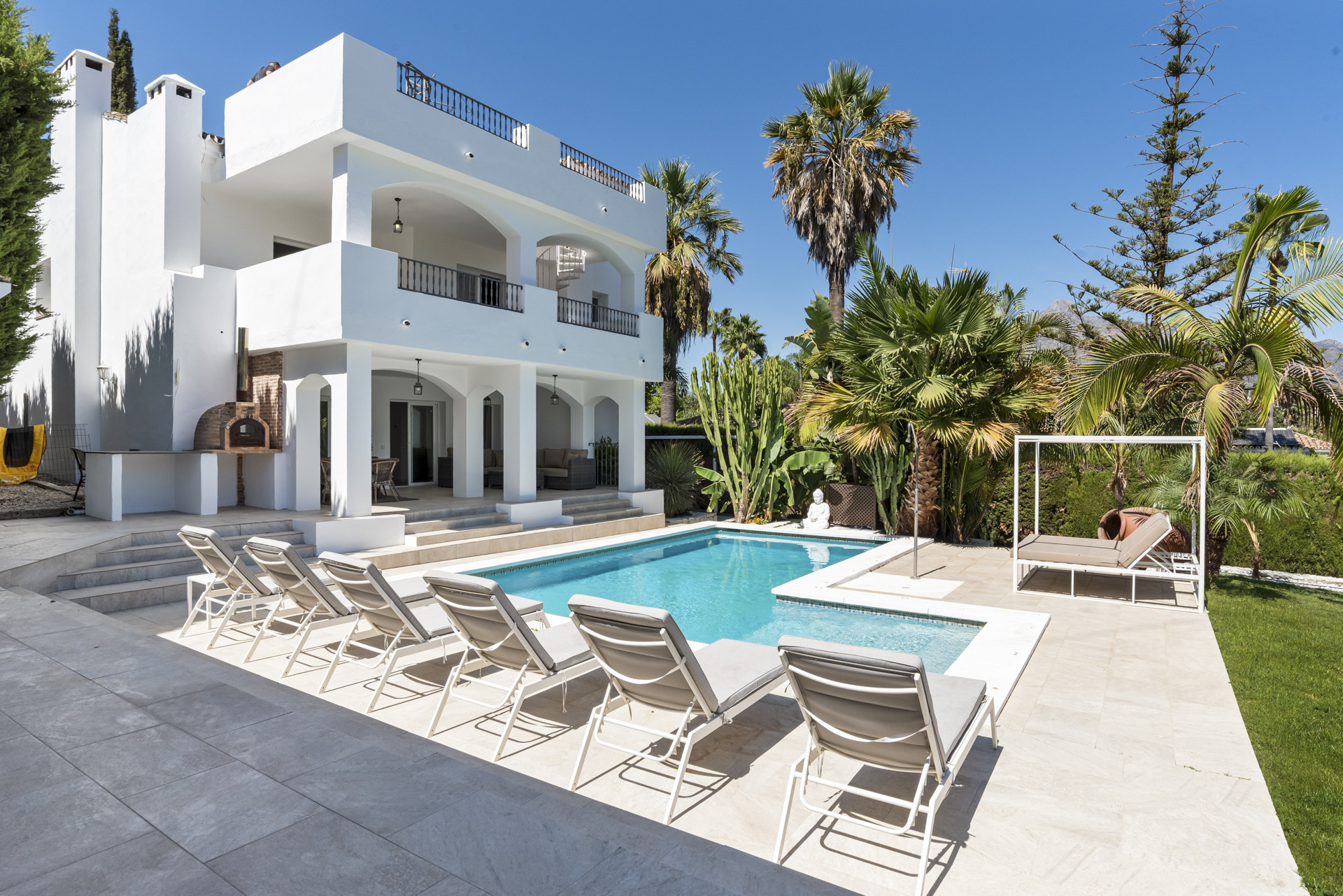 A white multi-story house viewed from the side, with a swimming pool, sun loungers, and tropical plants.