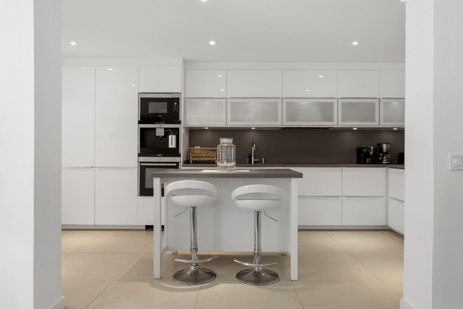Contemporary white kitchen with integrated appliances, light tile flooring, and a gray countertop island with two stools