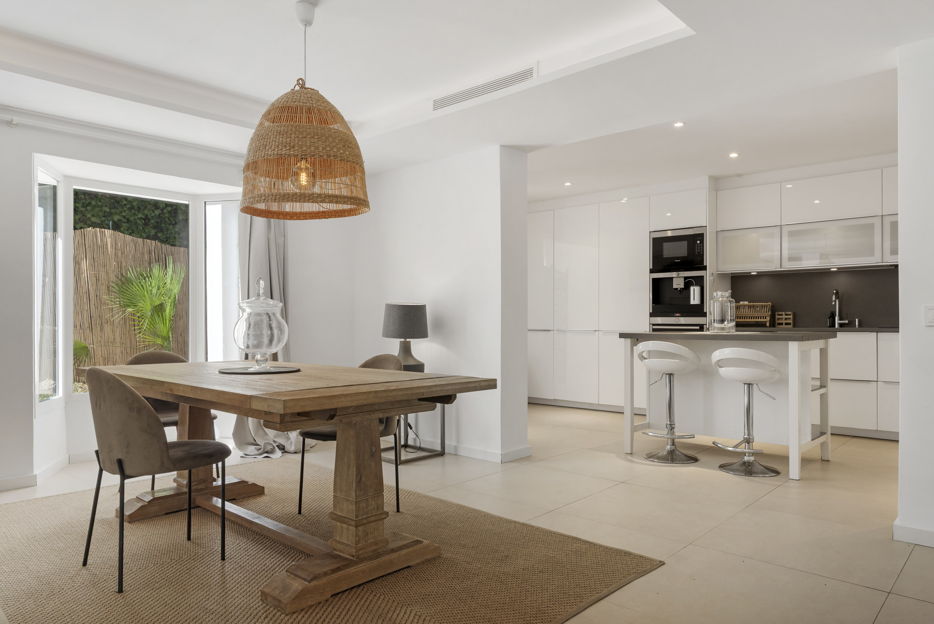 Dining area with a large wooden table, woven pendant light, and a modern kitchen with island in the background.