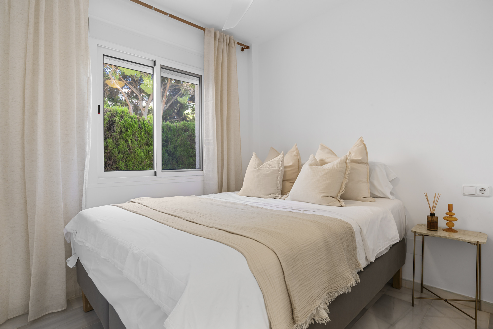 Bright bedroom featuring a bed with beige linens and a window showing green foliage outside.