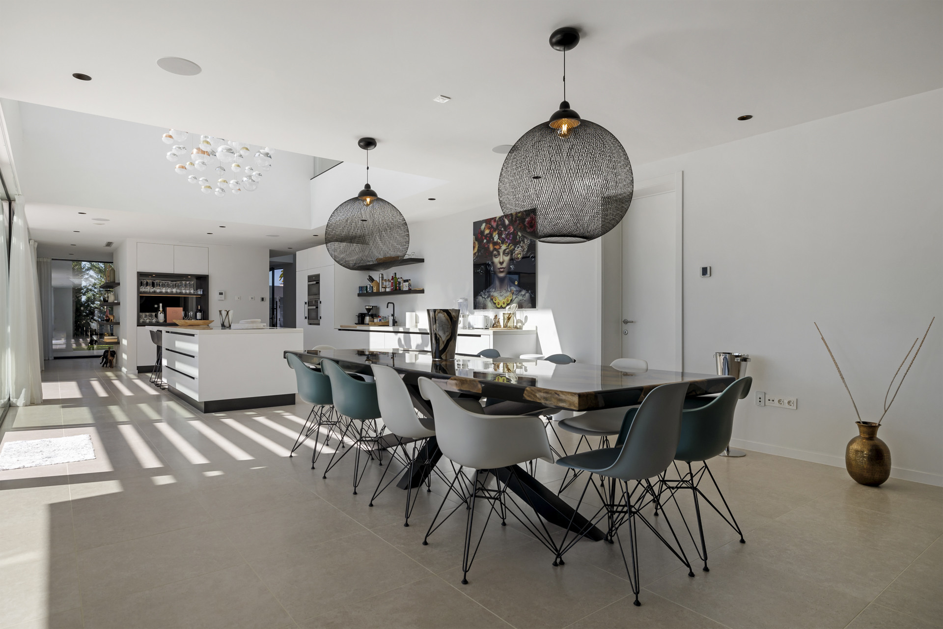Dining area with a long wooden table, modern chairs, three large black pendant lights, and kitchen beyond.