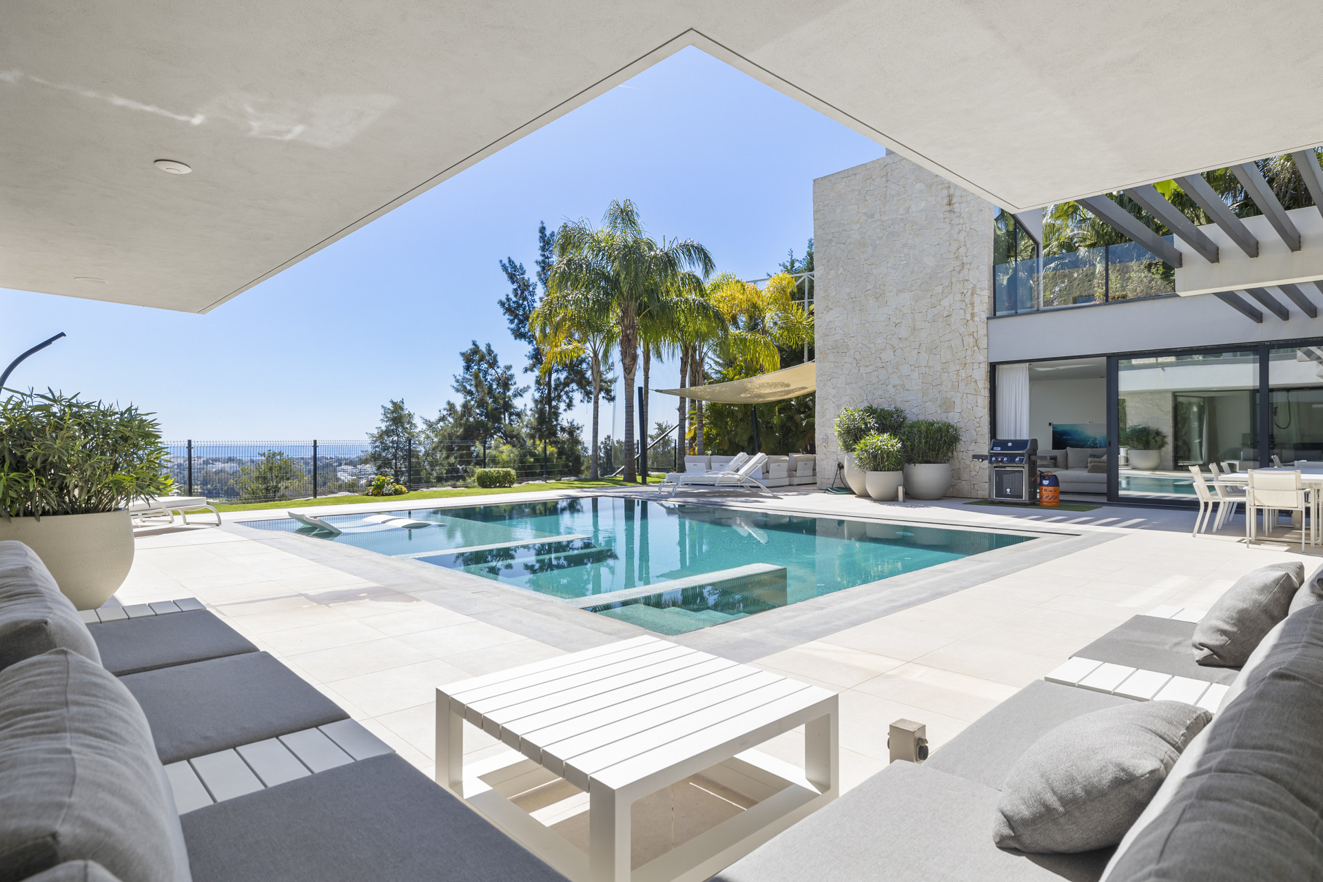 Outdoor patio with a white lounge set, swimming pool, and stone-clad exterior under a clear sky.