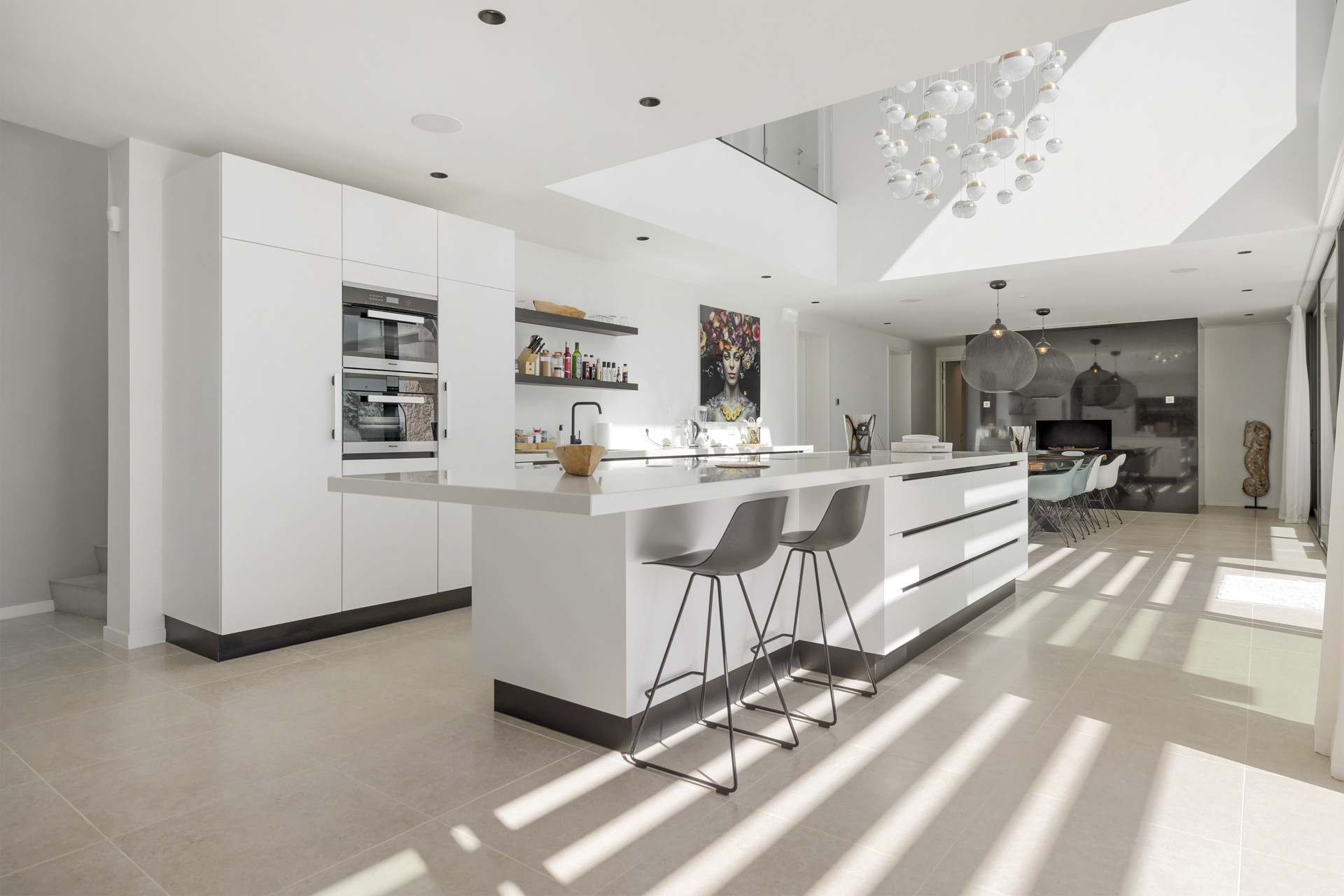 White modern kitchen with island and two barstools. Dining area is visible beyond with a large abstract artwork.