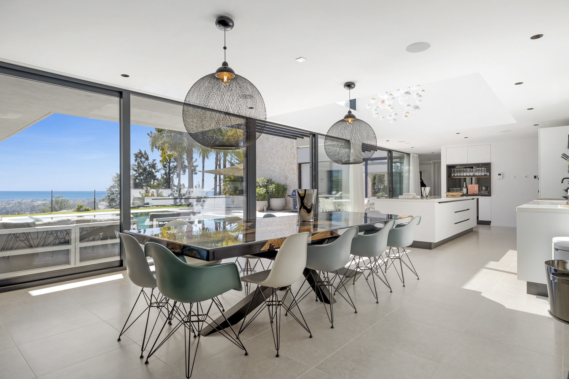 Modern dining room with large table and black pendant lights next to glass doors overlooking a pool and ocean view.
