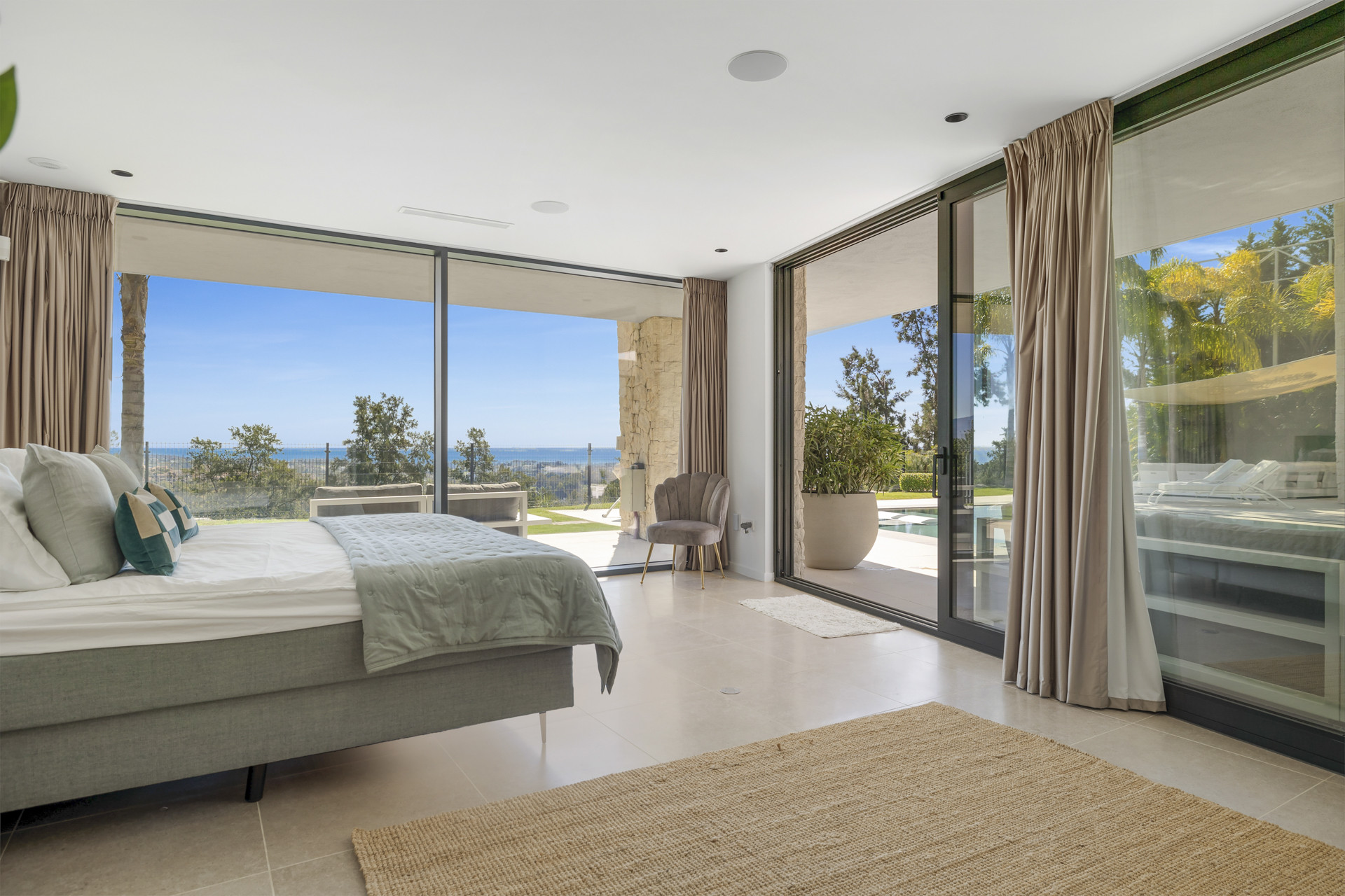 Bedroom with a large bed, full-height glass doors opening to a patio, pool, and distant ocean view.