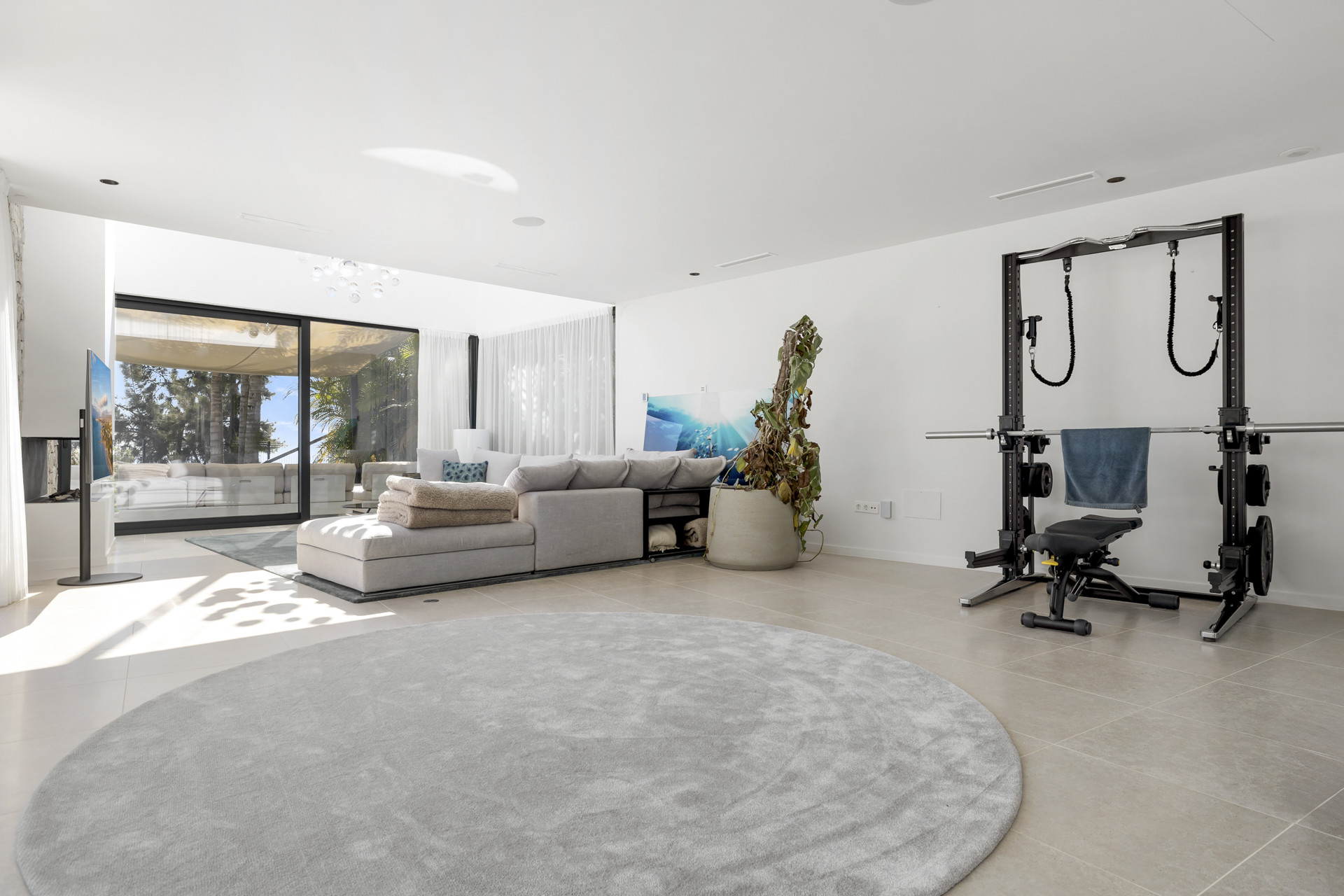 Living room with gray sectional sofa, large rug, and gym equipment in one corner. Glass doors lead outside.