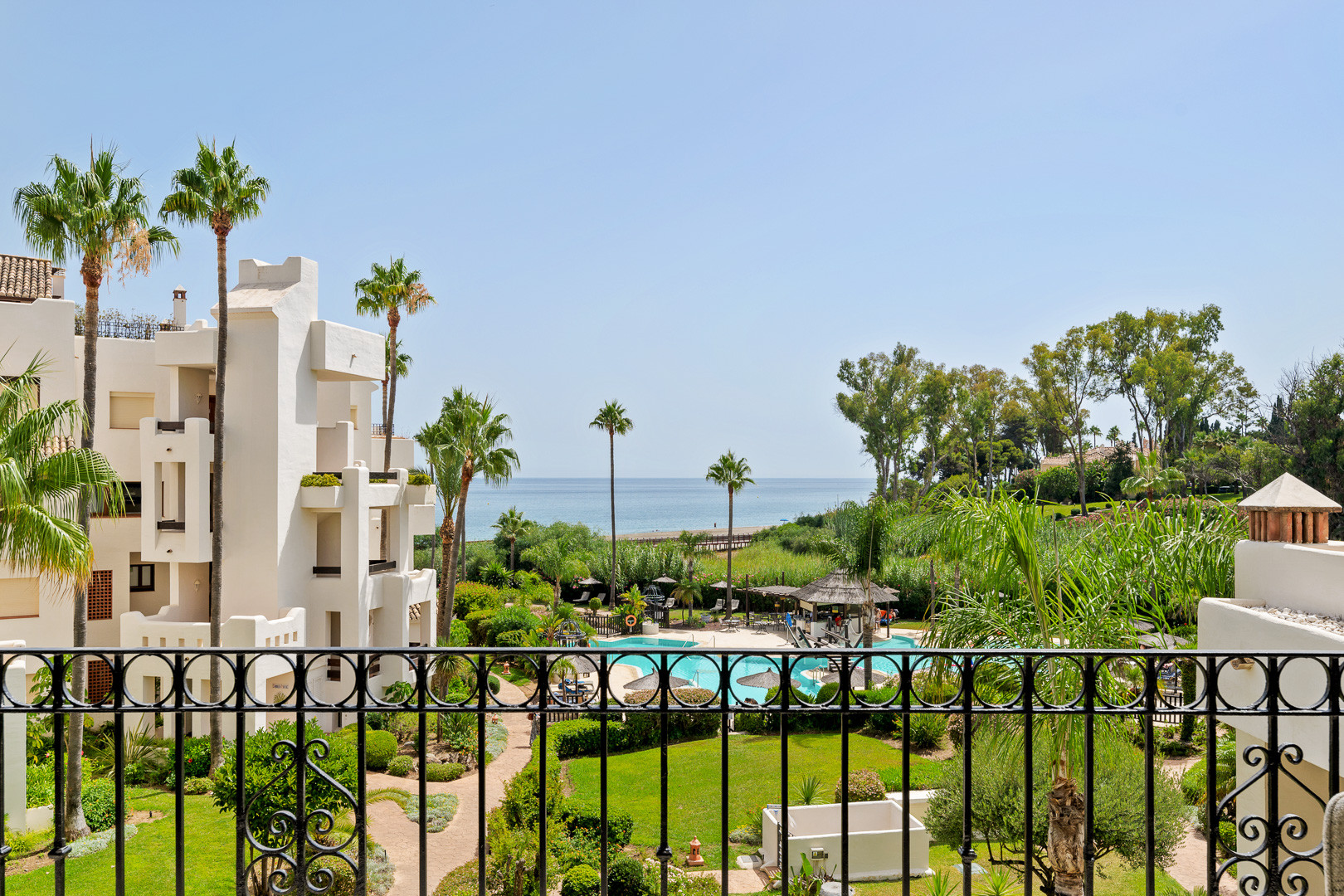 Balcony view over a black railing showing a resort pool area, palm trees, buildings, and the sea.
