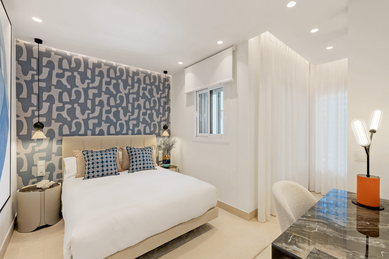 Bedroom with a patterned blue accent wall, double bed, window, and a desk in the corner.