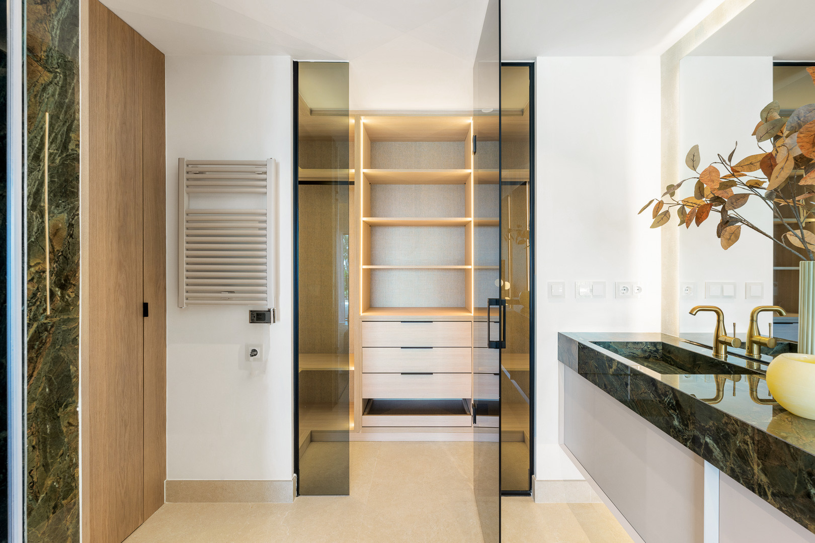 Bathroom vanity with marble countertop and gold faucets, next to a glass-enclosed walk-in closet.